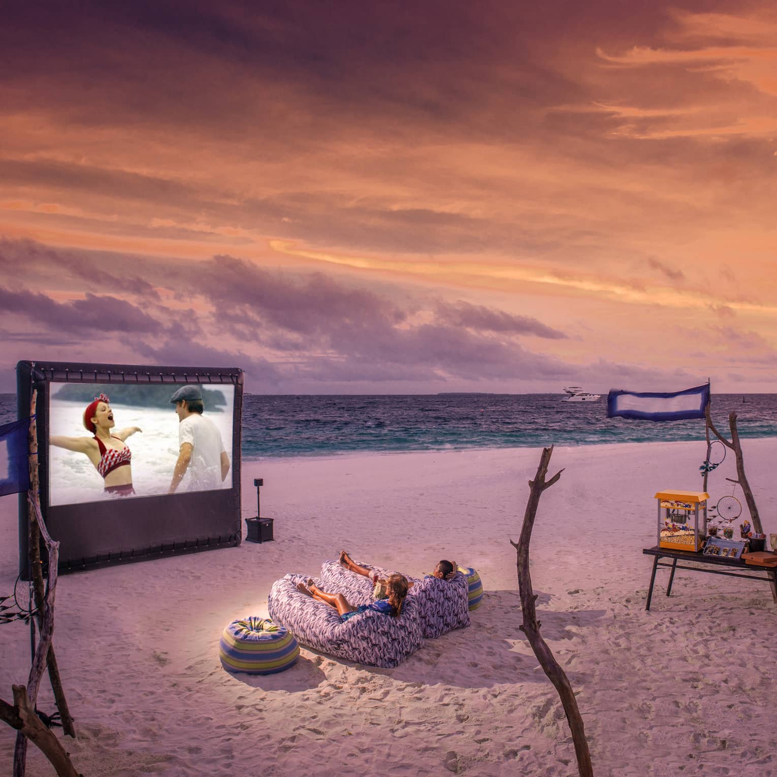 ,Two people in beanbag chairs enjoy a movie night on the beach at dusk, popcorn stand on sand behind them