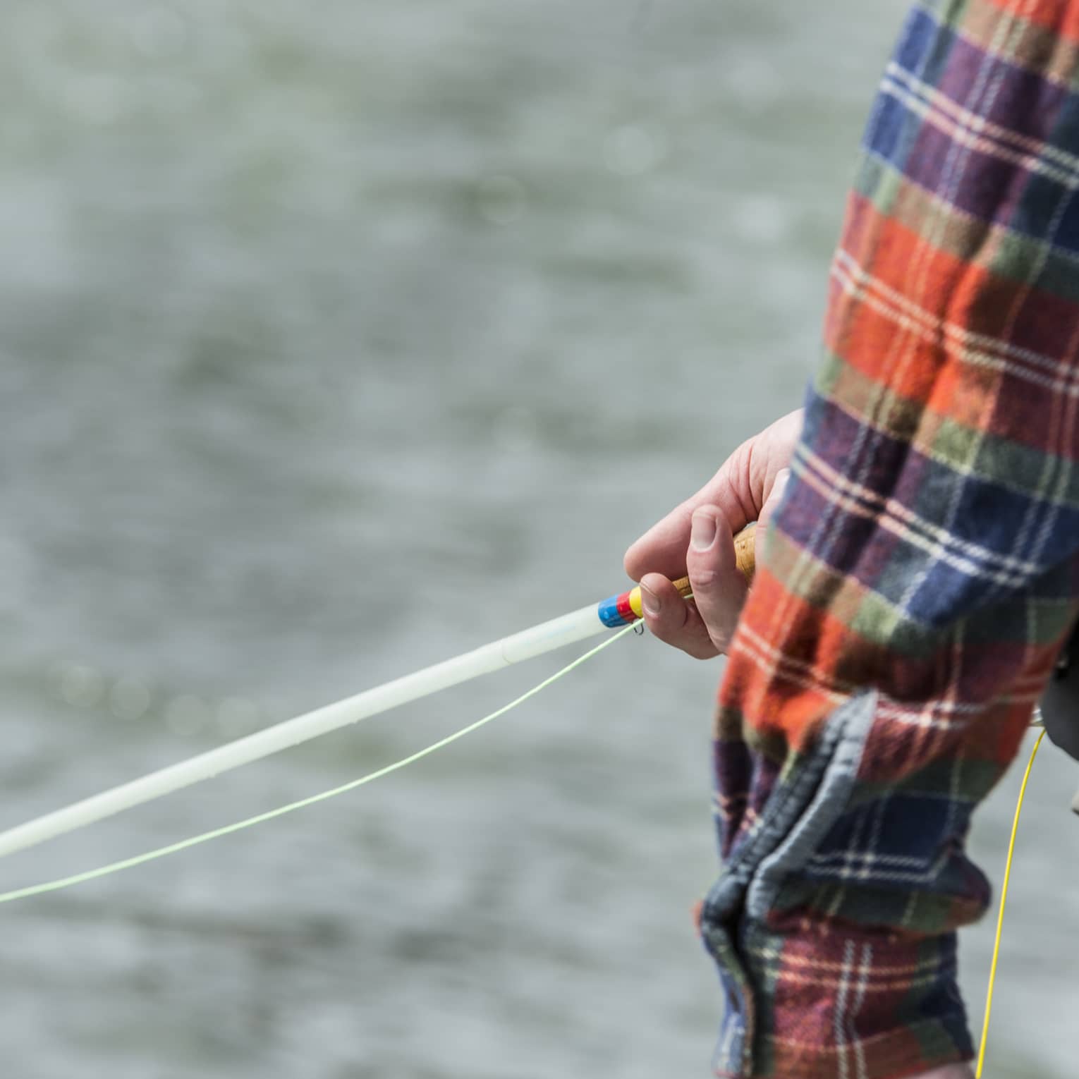 Against a background of rippling silver water, a plaid sleeve partly reveals a hand gently holding a colourful fishing rod.