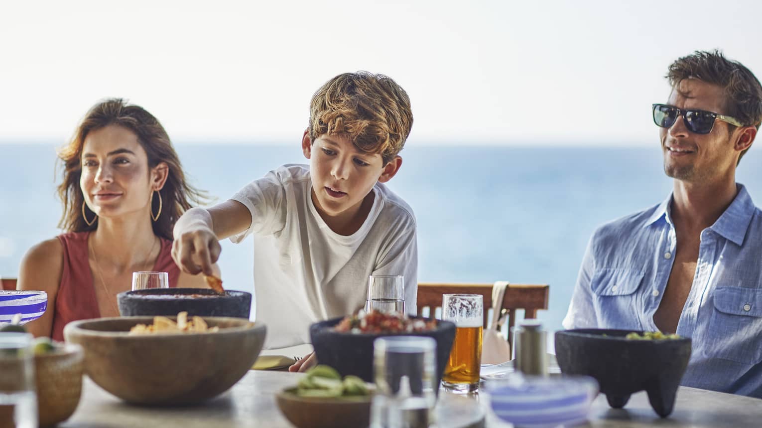 A young boy reaches for a chip bowl on table as a woman and man sit on either side, ocean in the background