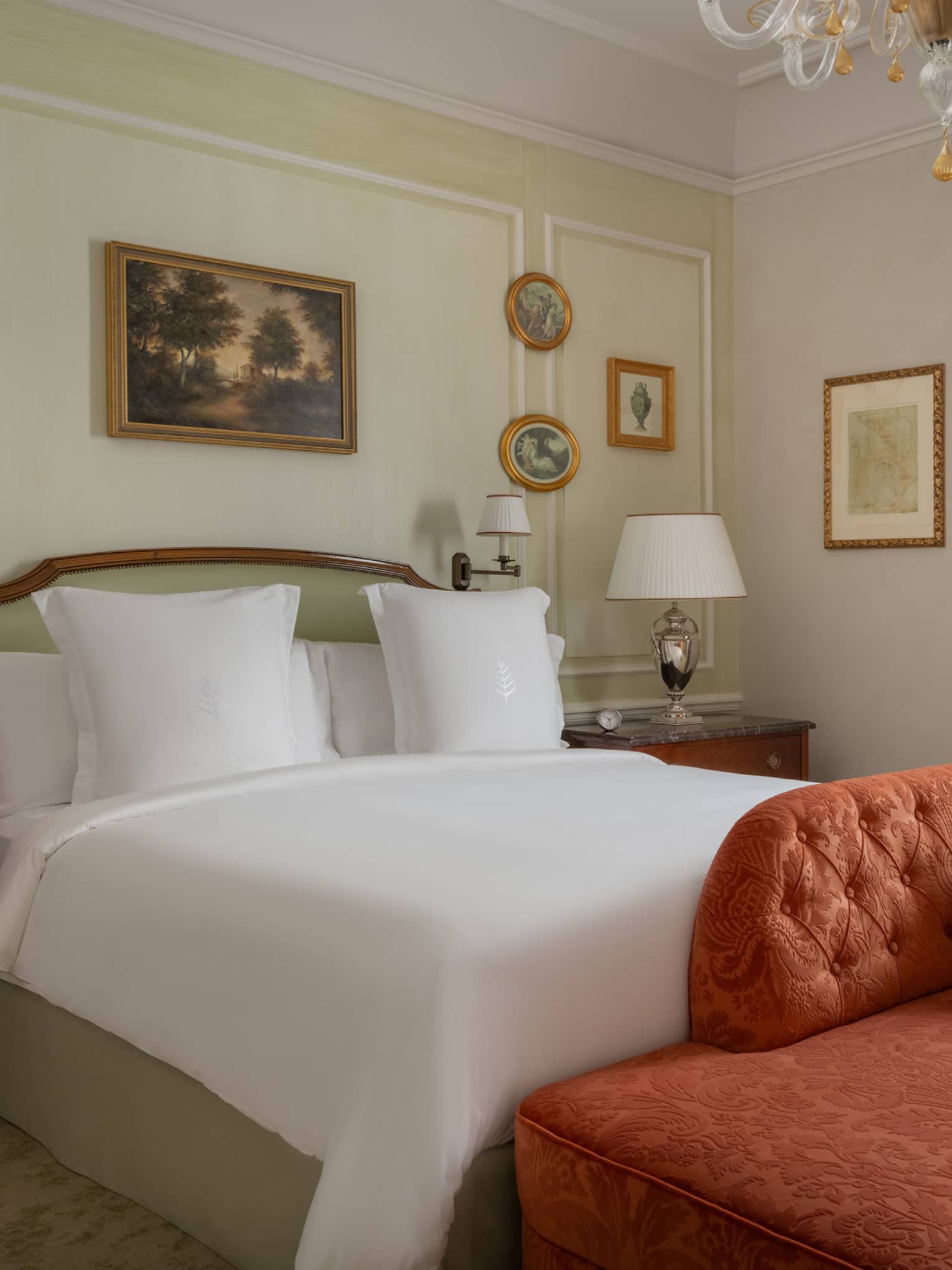 Elegant hotel room with king bed, orange chaise at the foot of the bed, chandelier, window with curtains