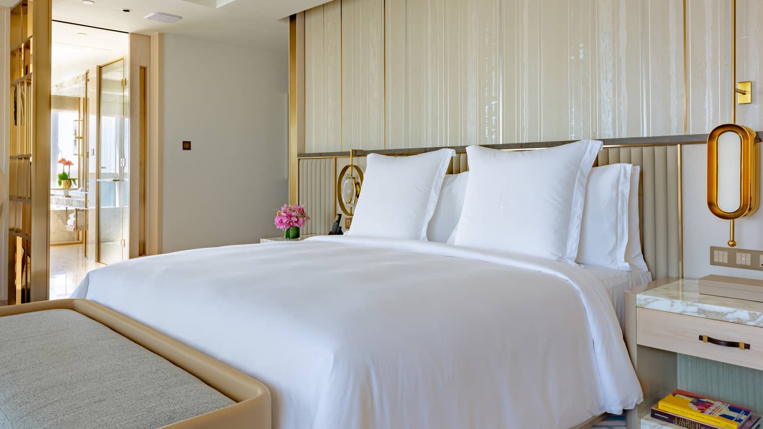 A perfectly-made bed in a contemporary bedroom leading into gold-trimmed bathroom.