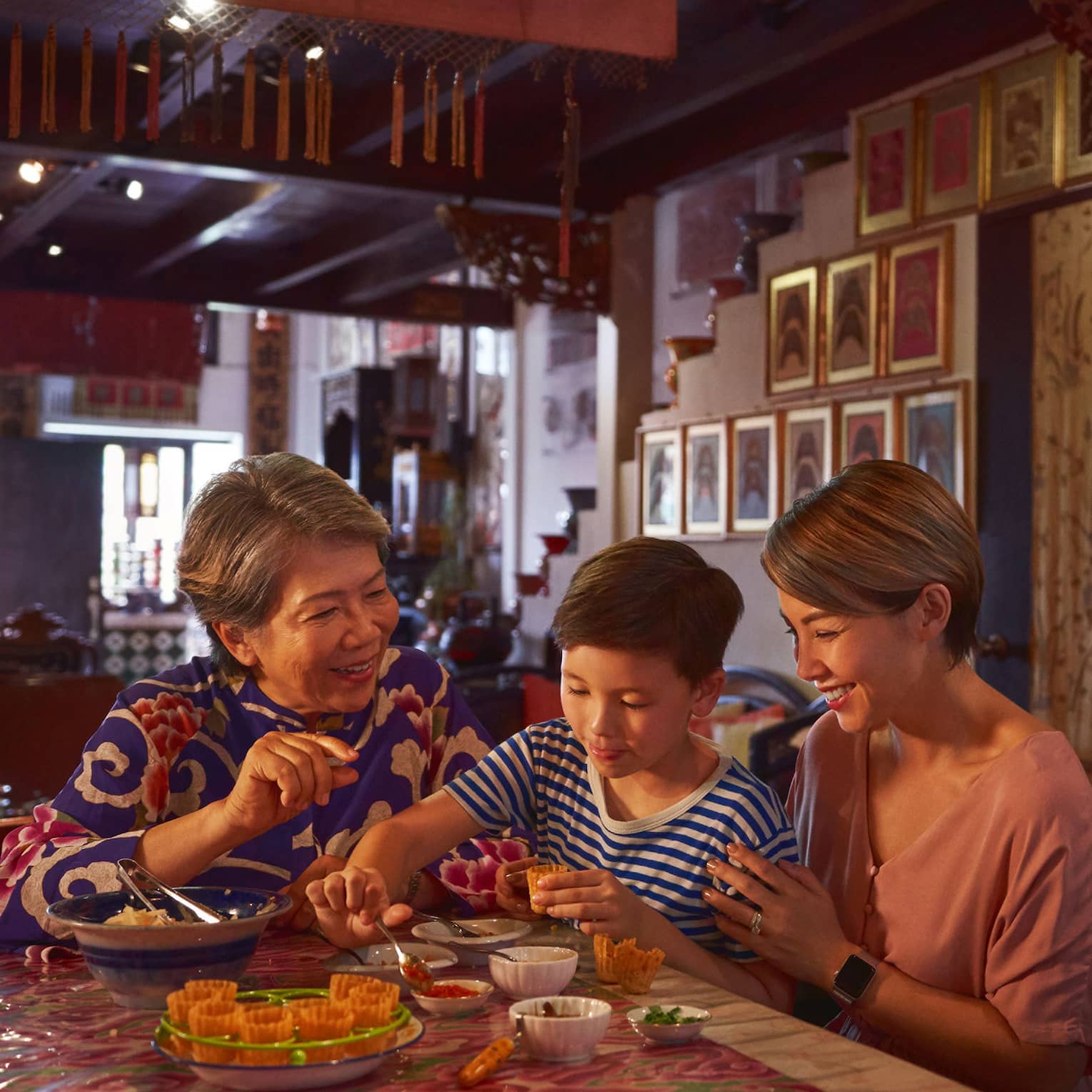 A family sits in a richly decorated, dimly-lit room at a table with various bowls as a child scoops jam into a pastry shell.
