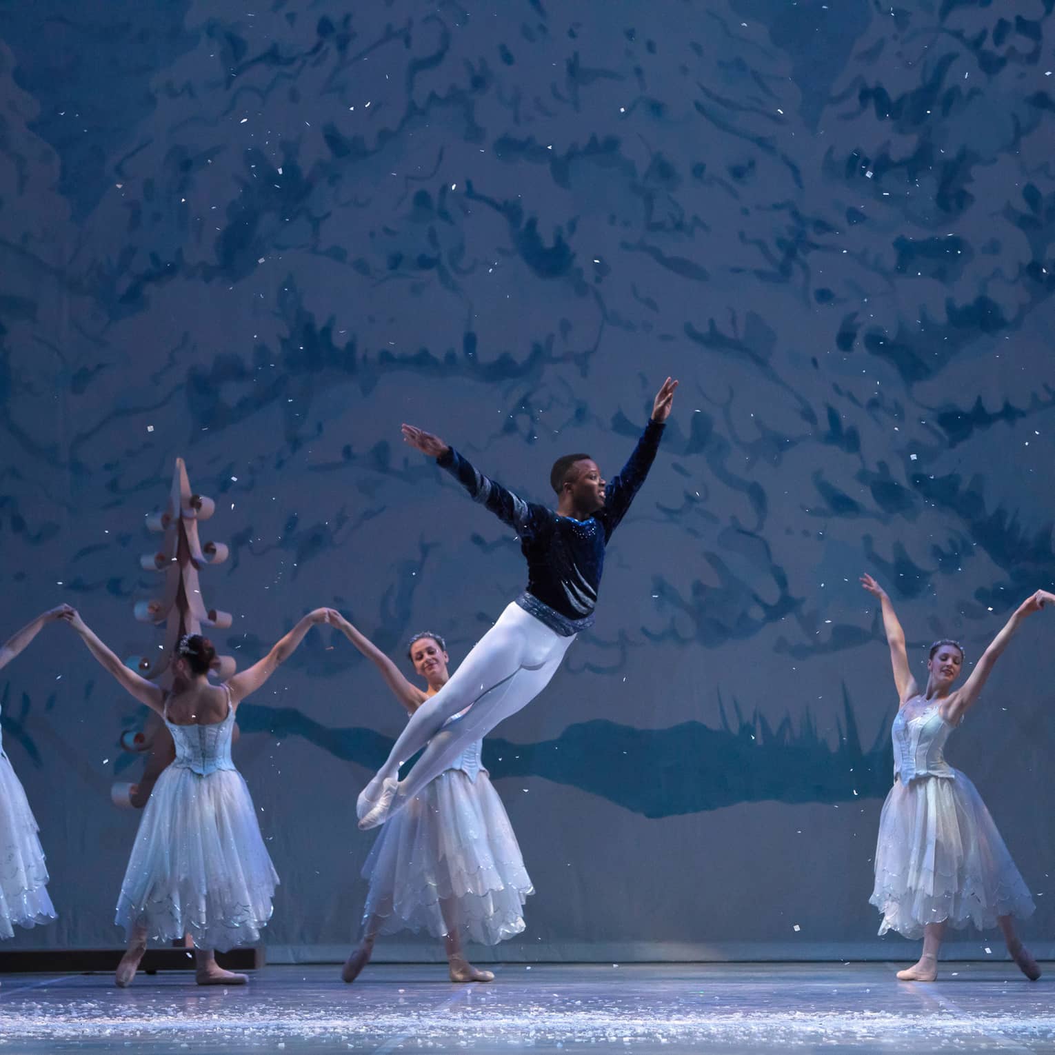 Ballerinas on stage in long ice-blue tutus dance before a snowy painted backdrop as a dancer wearing a leotard leaps grandly.