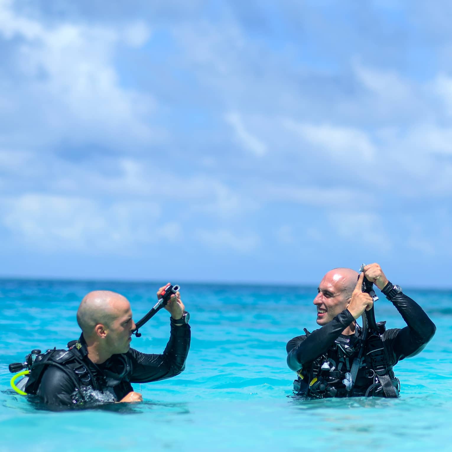 Scuba instructor showing mouthpiece to guest as they stand wearing scuba gear, chest-deep in azure water under fluffy clouds.