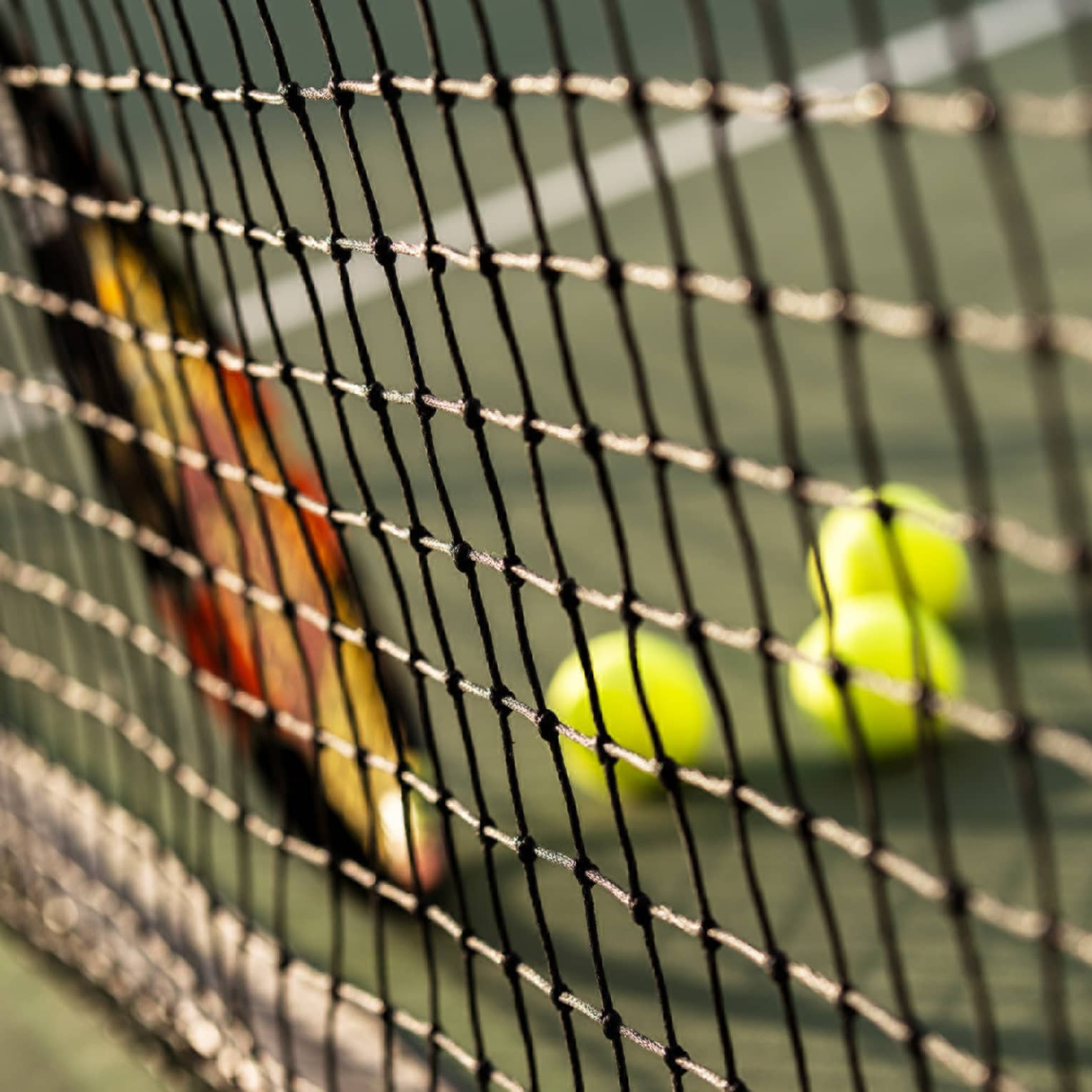 Close-up of tennis racket against net, three green tennis balls on ground