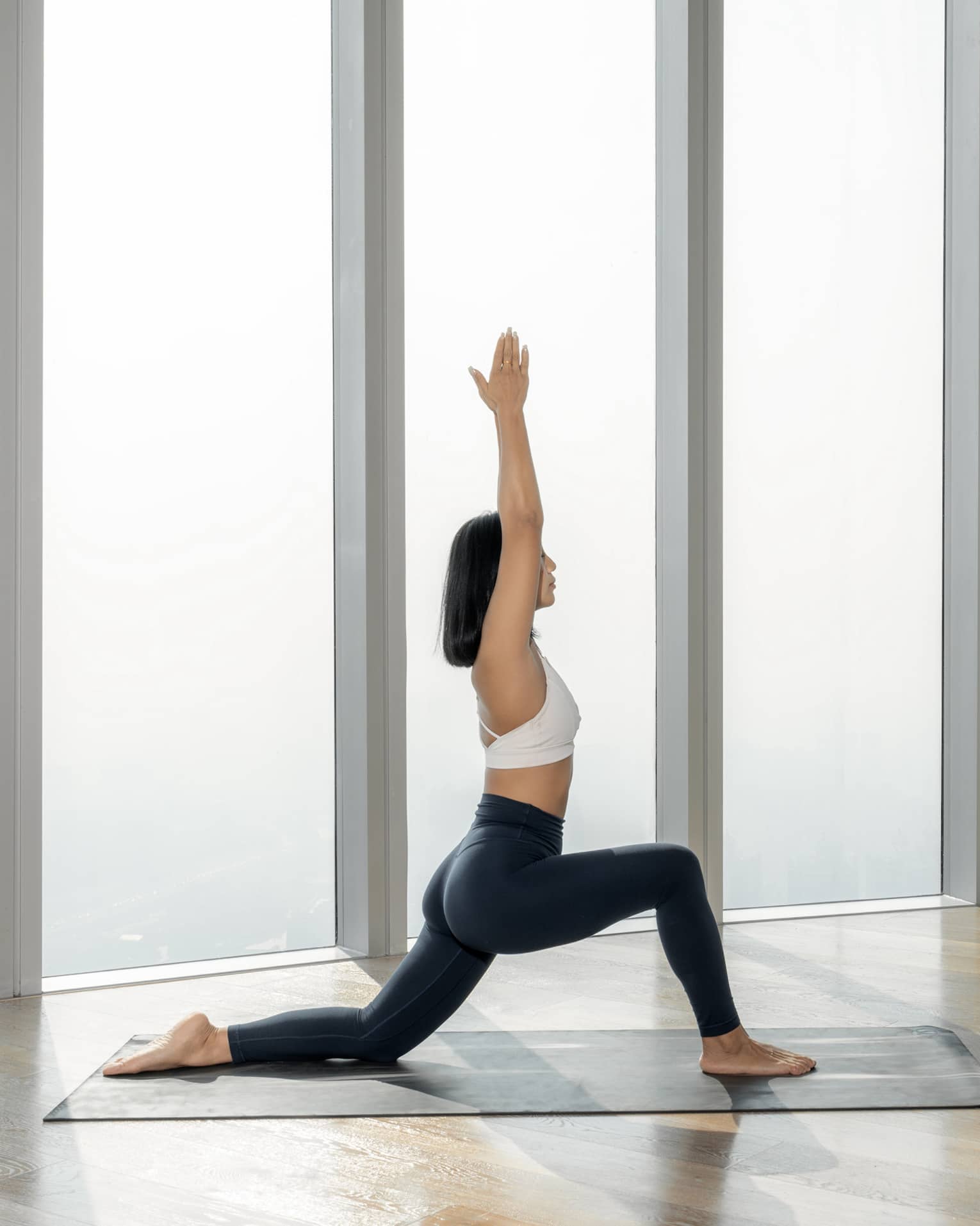 Woman lunges on yoga mat with hands above her head, city draped in fog visible out of the floor-to-ceiling window