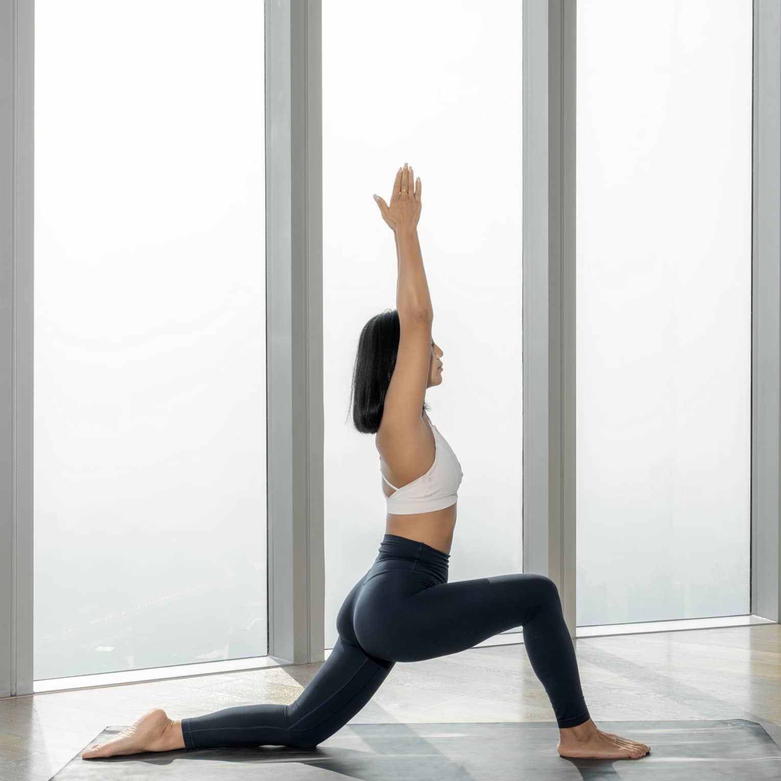 Woman lunges on yoga mat with hands above her head, city draped in fog visible out of the floor-to-ceiling window