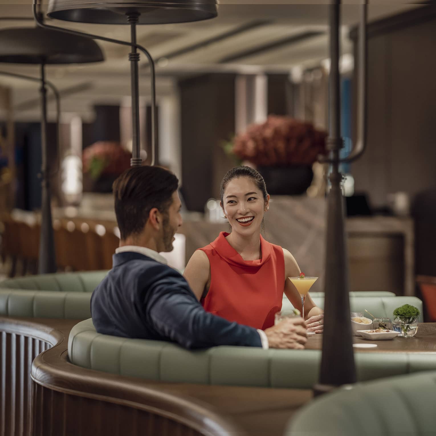 A smiling couple sit at a circular booth in an empty, dimly lit restaurant with cocktails and appetizers on their table.