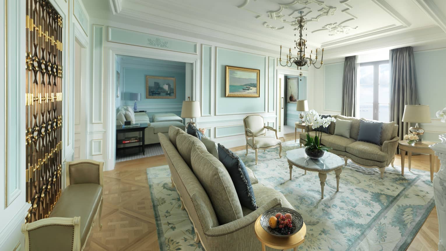 Presidential Suite sunny living room with two elegant sofas, coffee table under small chandelier by window, marble fireplace