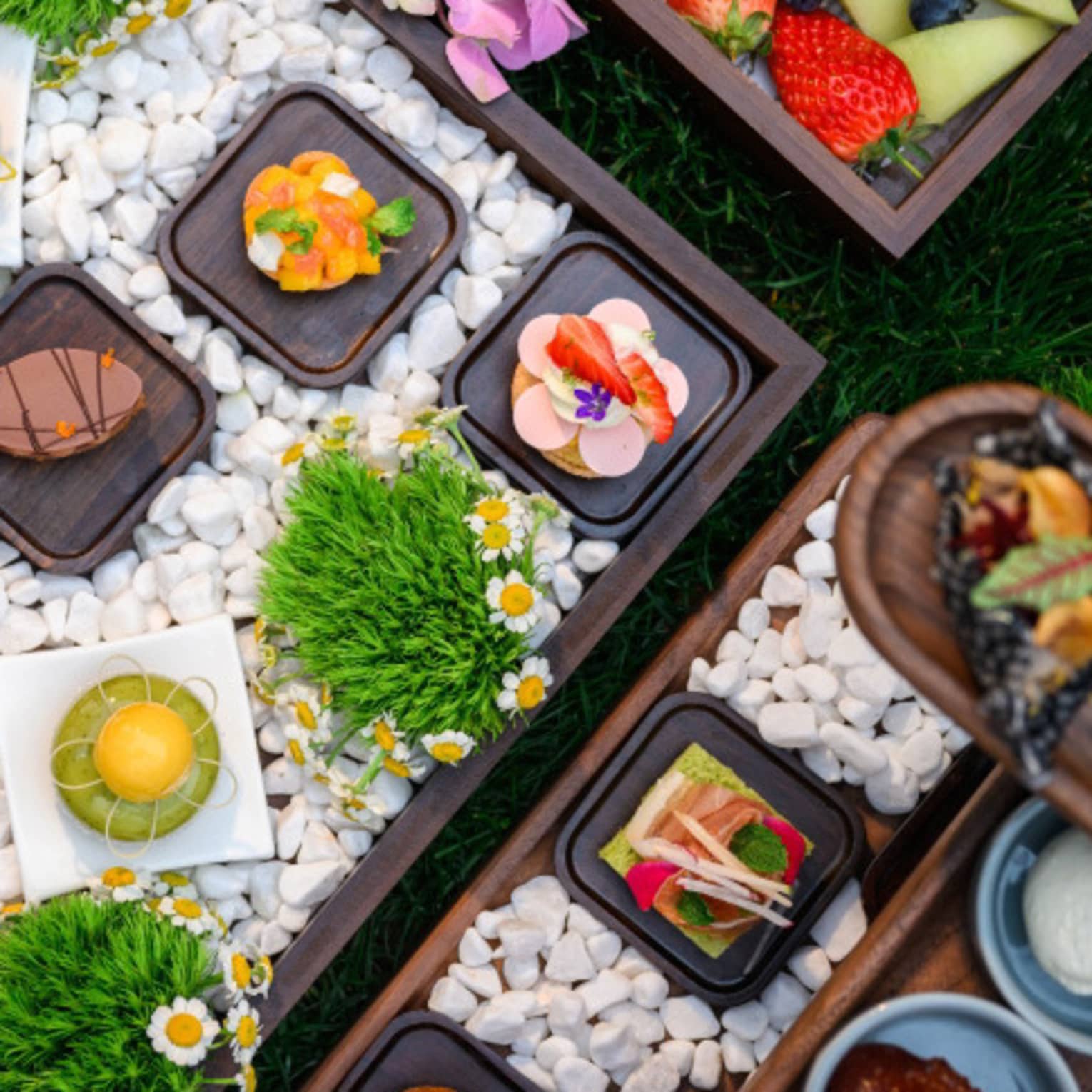 Wooden boxes laden with stunning, colourful, geometric individual tea delicacies and decorations atop pearly white pebbles.