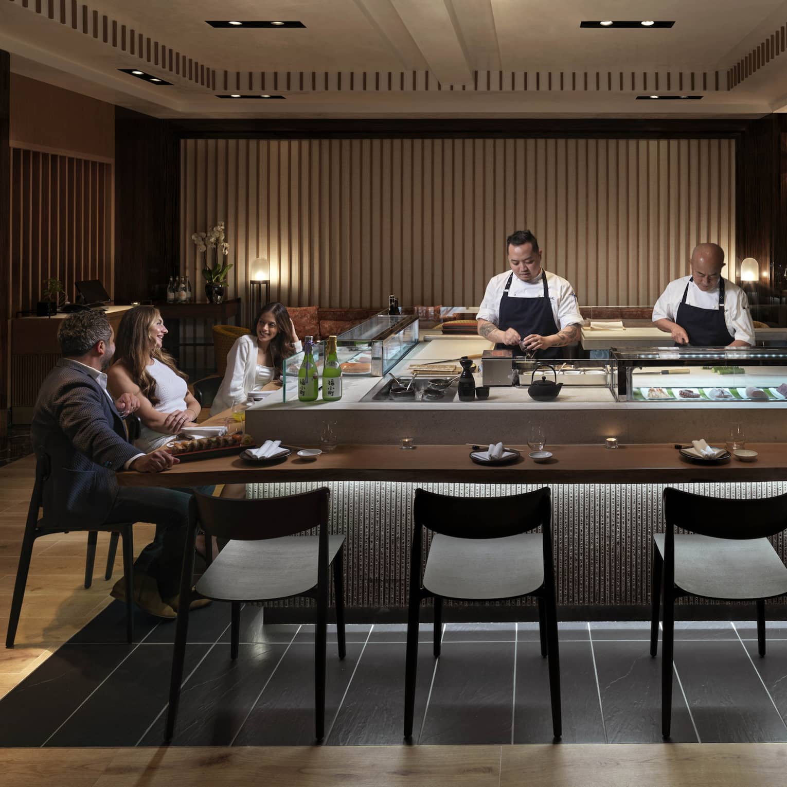 Three guests sit at a sushi counter, engrossed in conversation, while two chefs behind the counter carefully prepare food.