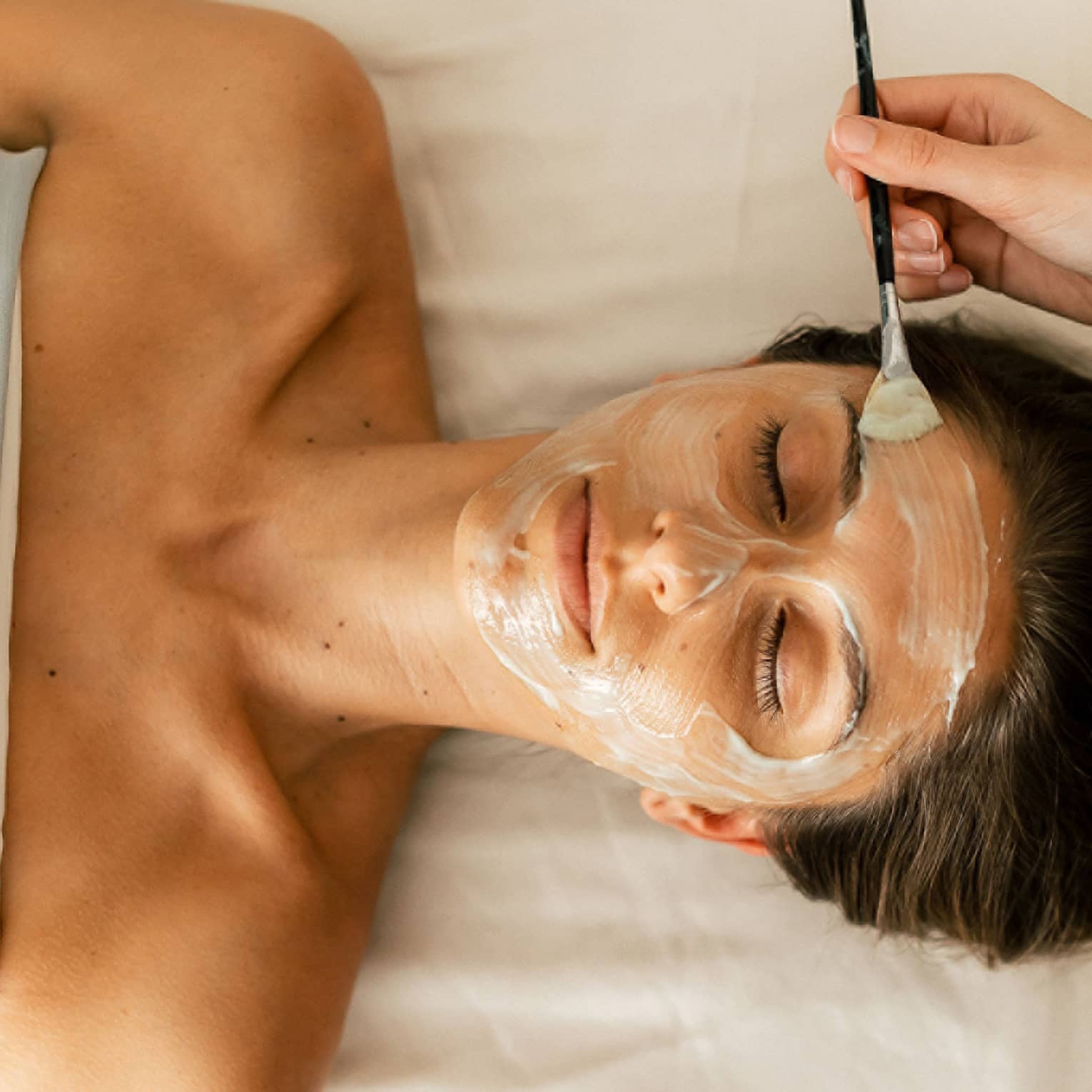A person holding a small brush applies a white face mask treatment onto a woman's face