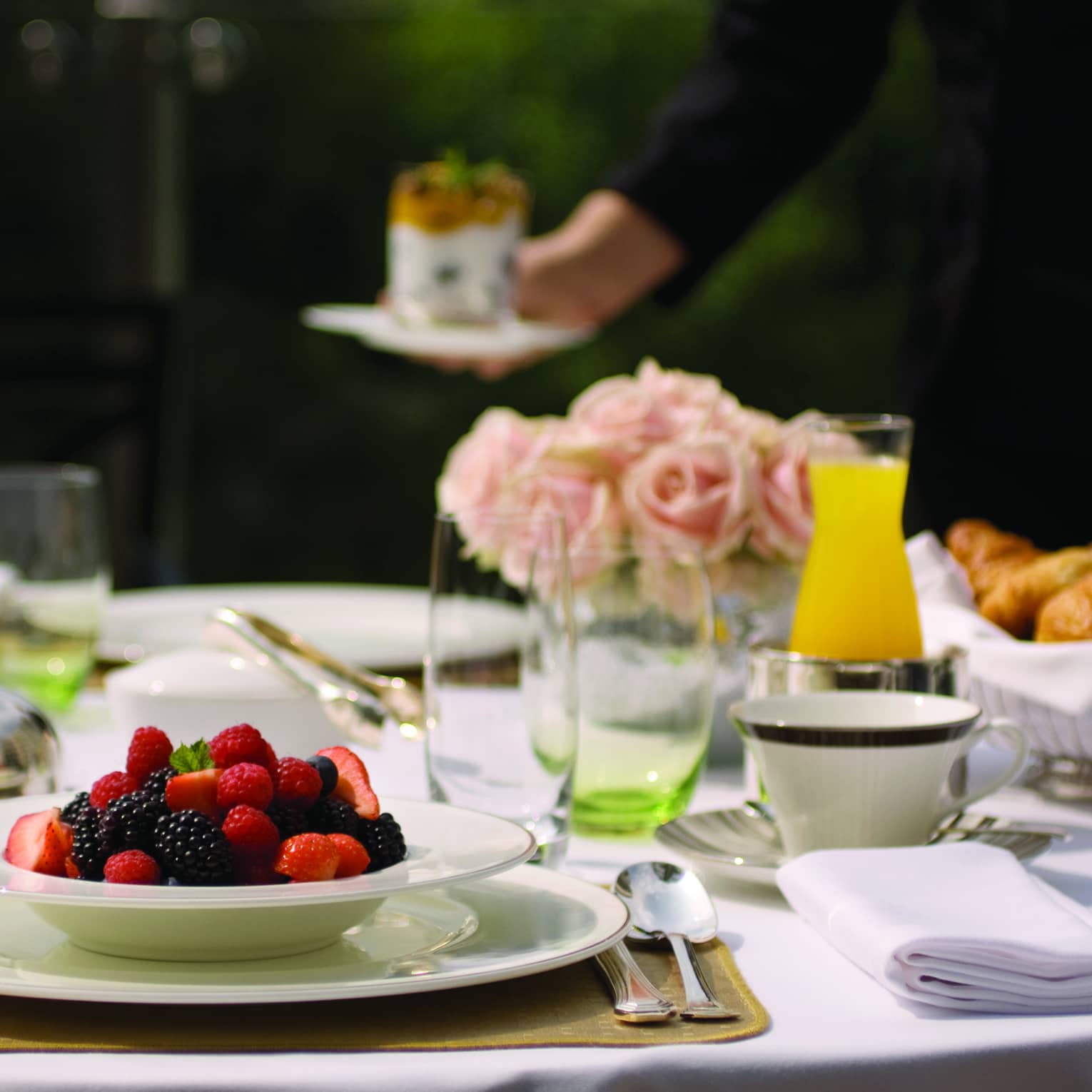 White bowl filled with fresh strawberries, blueberries and blackberries on table with glasses, orange juice, pink roses