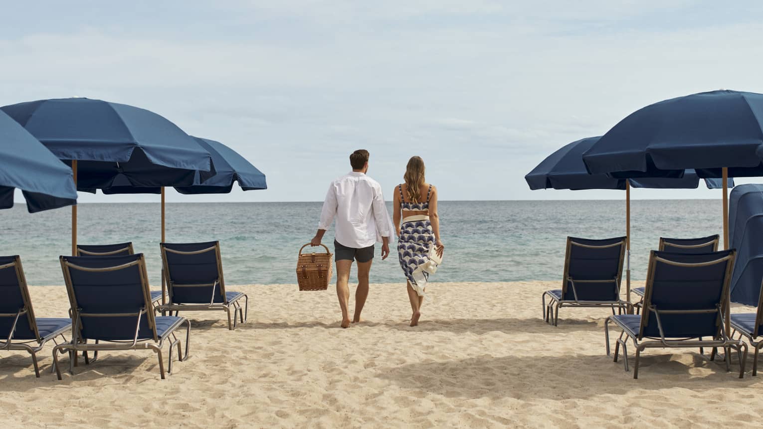 A man and woman walking between blue lounge chairs and umbrellas on sand towards the ocean.