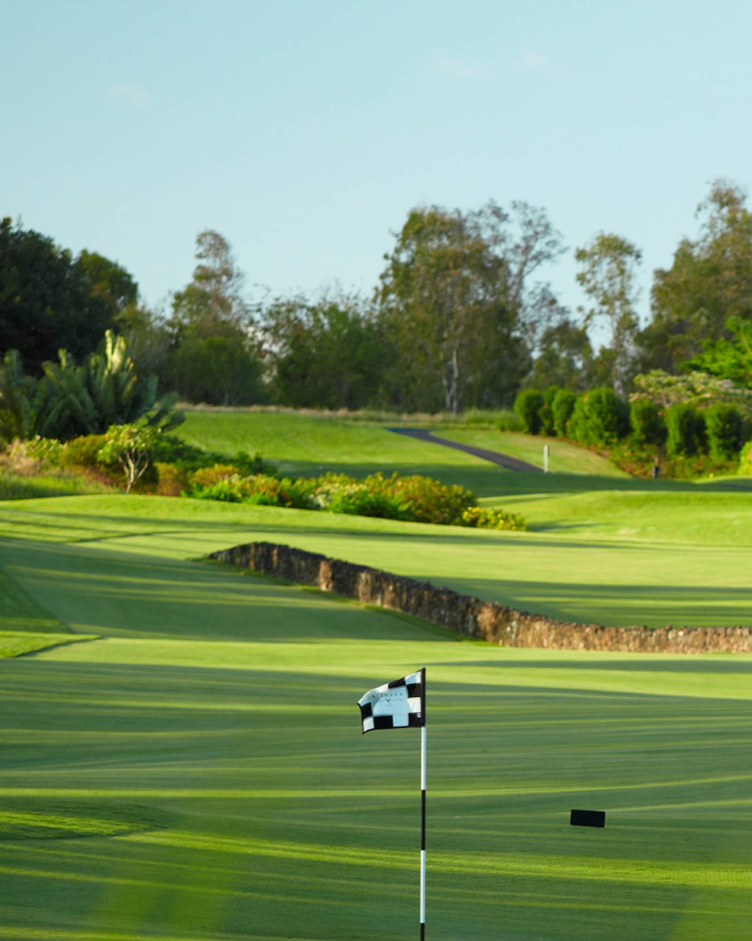 Man with golf club walks toward flag on golf course green near stone fence