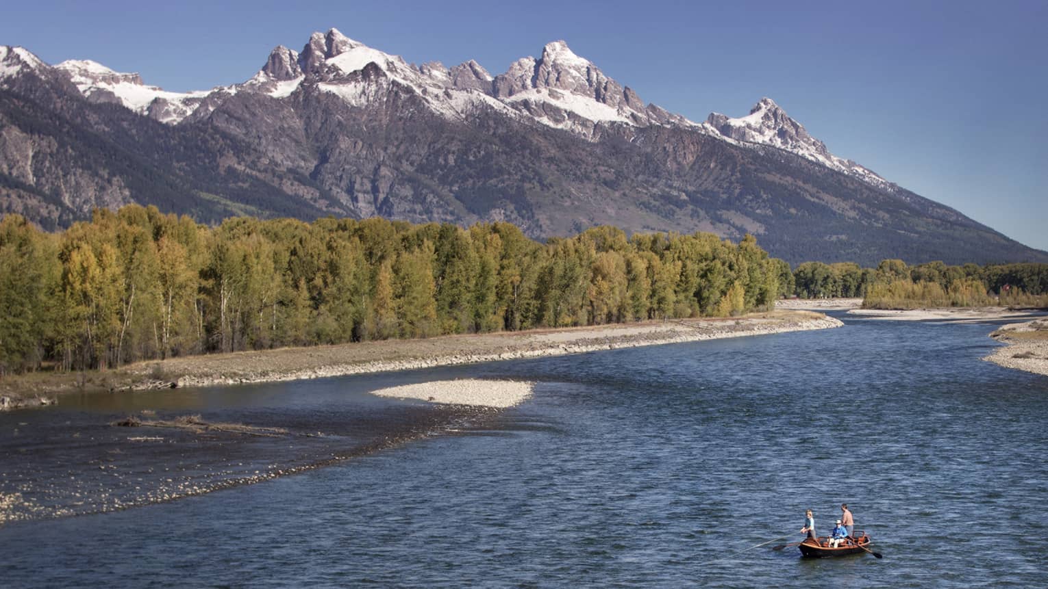 People fishing in boat on wide river by rocky shore, green trees, mountains