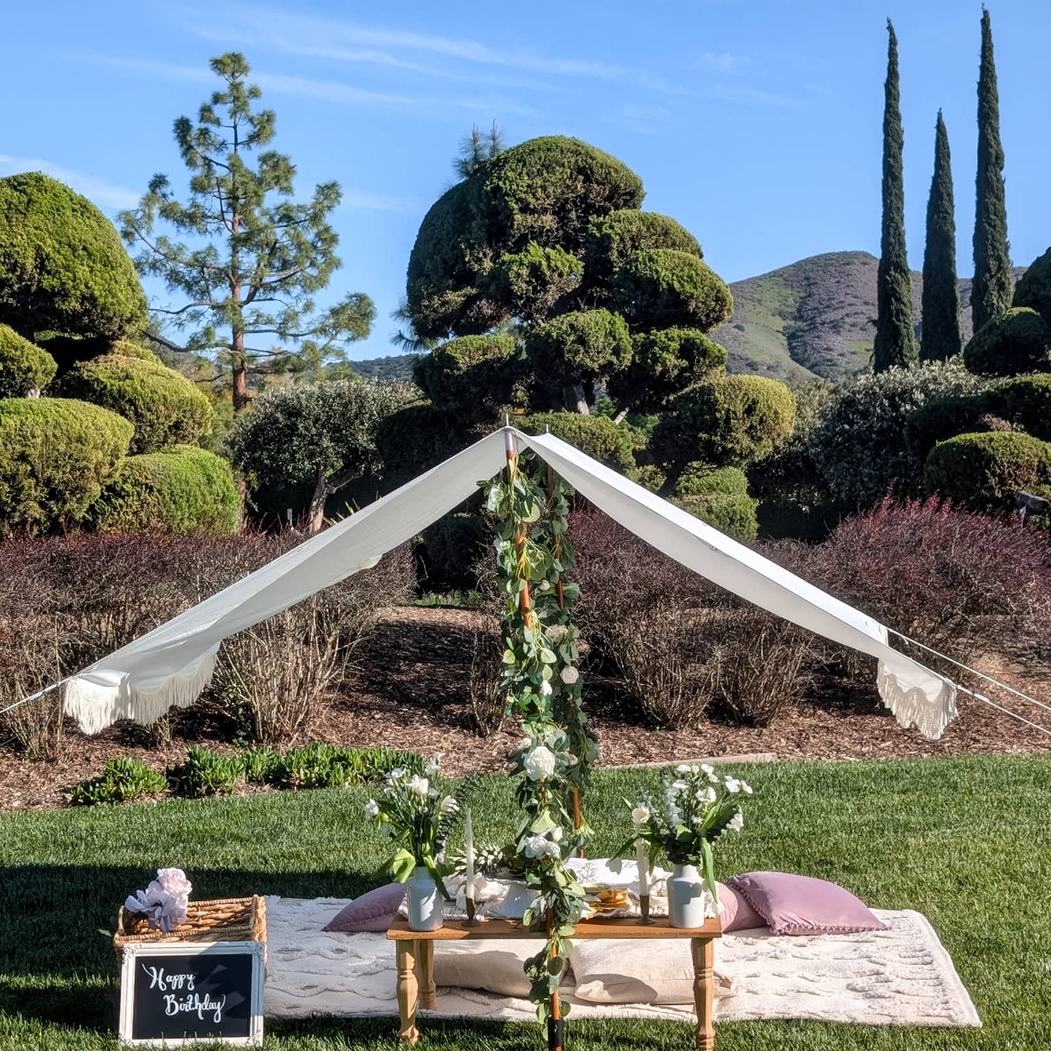 Picnic set up on a lawn with blanket, pillows, small table, flower garlands and white canopy