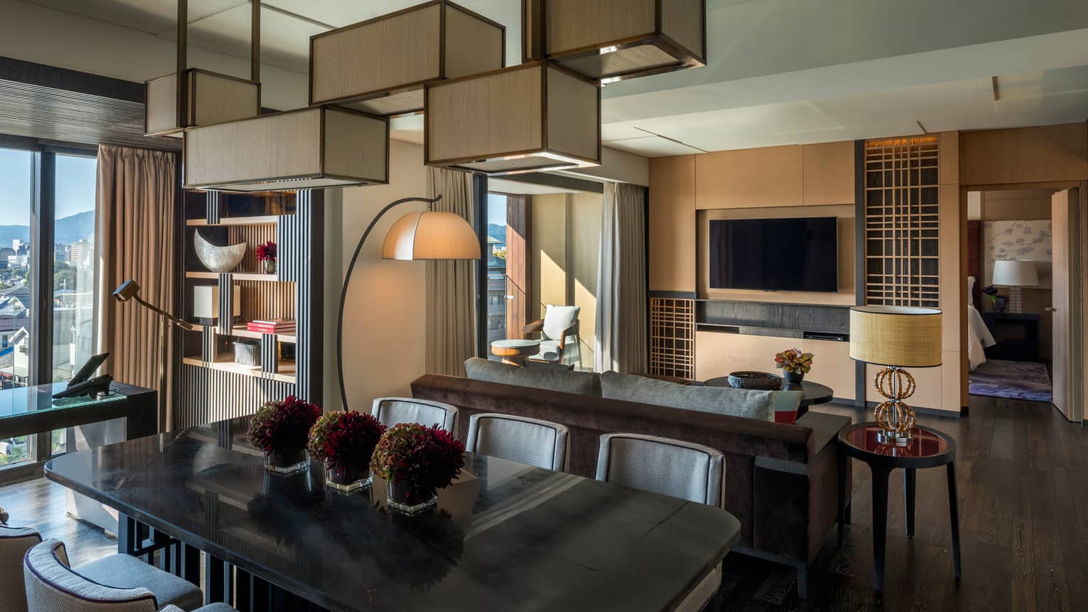 Residential suite with dining table, sofa, wall-mounted TV, floor lamp, bookshelf, desk with leather chair and floor-to-ceiling views of the city skyline