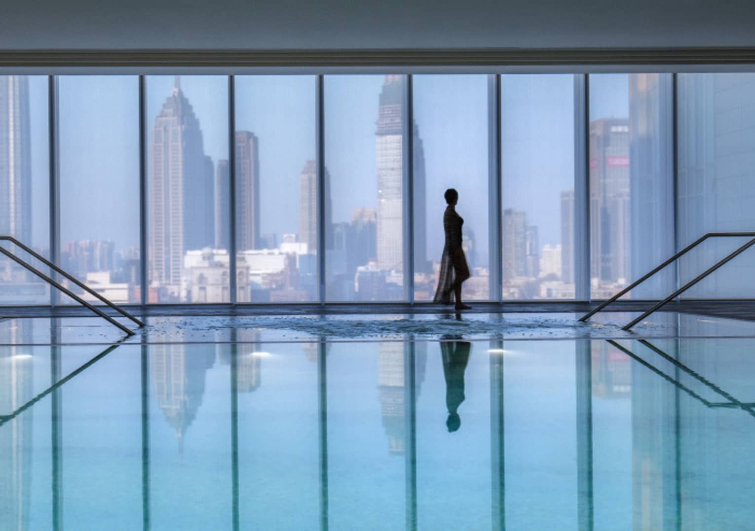 Silhouette of woman walking by blue indoor swimming pool, floor-to-ceiling window