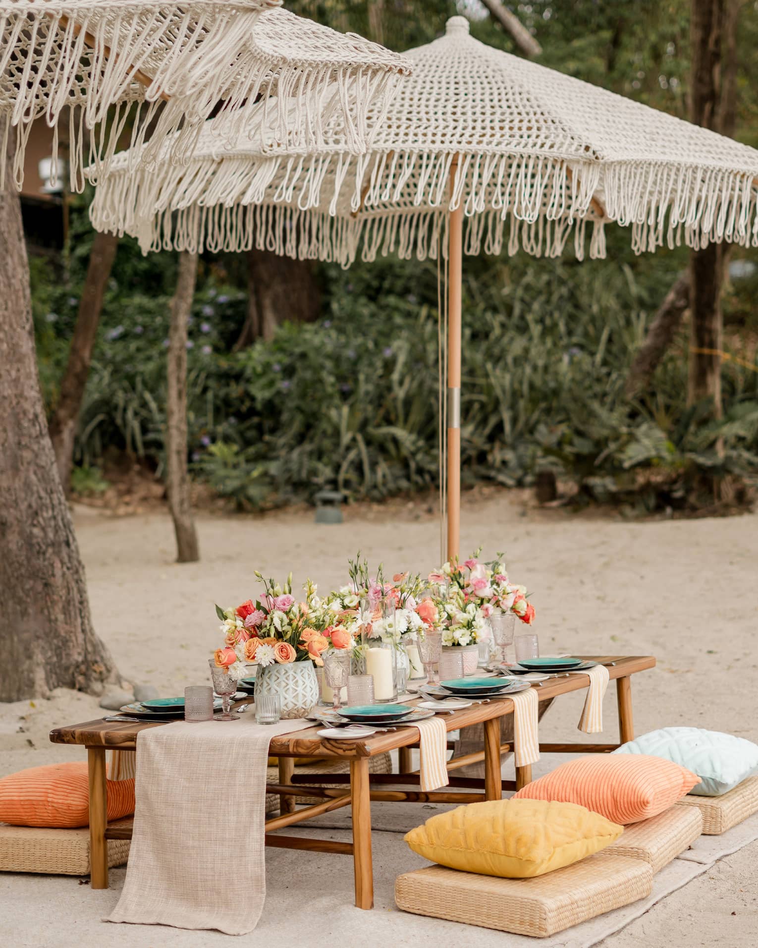 Wooden dining table set up with floral centrepieces and candles beneath an umbrella on a beach
