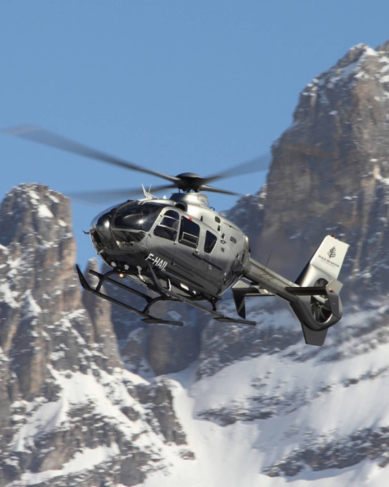 Silver helicopter ascending beside snowy, rocky mountain peaks