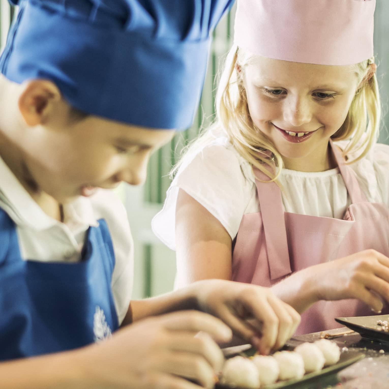 Three children in chef hats, aprons roll pastries at counter 
