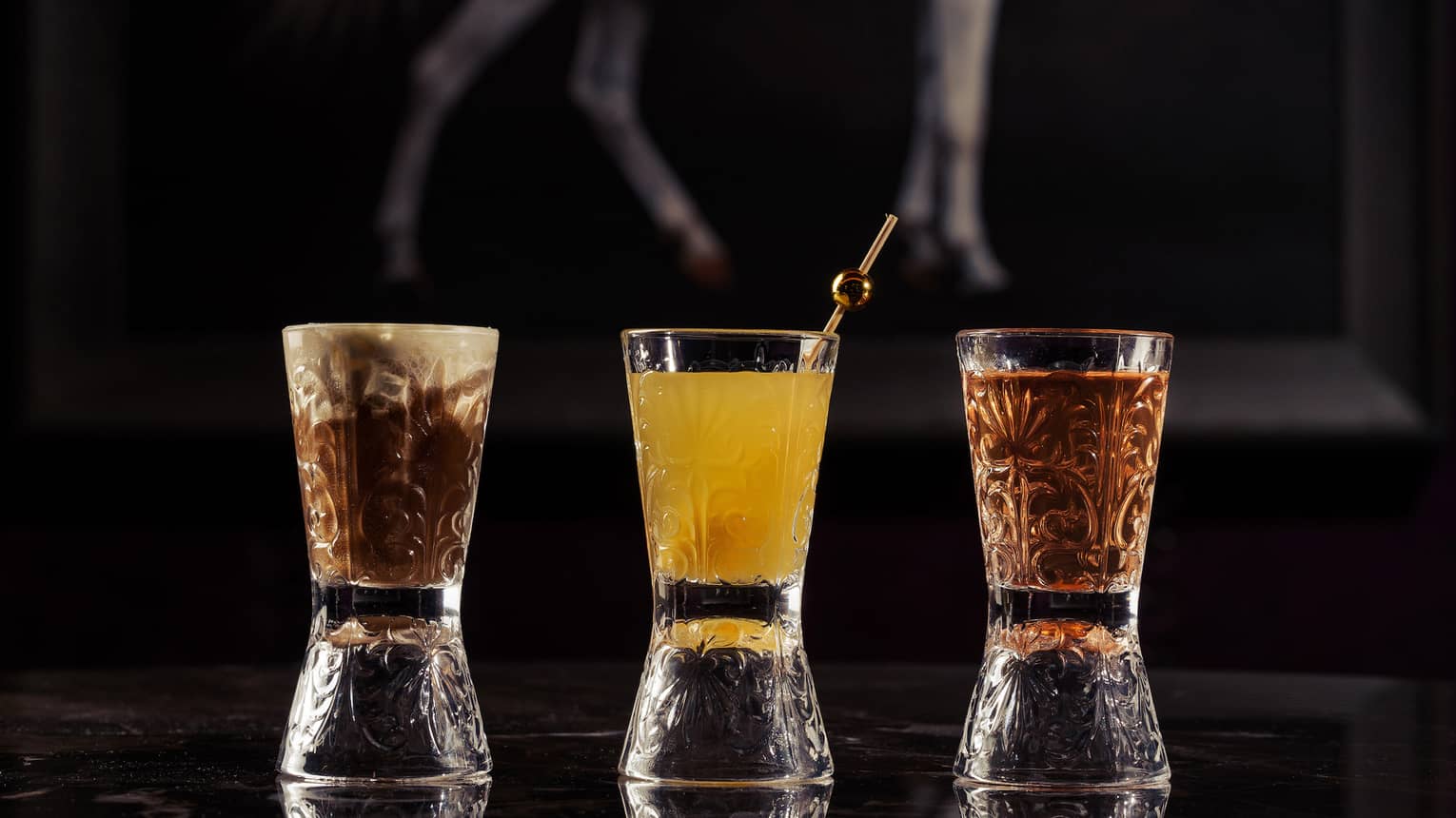 Three shot glasses in a row, the one on the far left holding a brown beverage, the one in the middle holding a yellow beverage and the one on the right holding a carmel-colored beverage, are placed on a dark table in front of a painting of a white horse