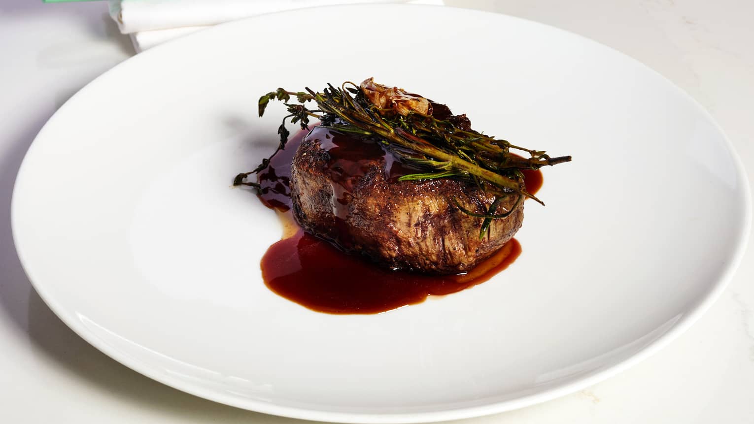 A white plate with a cooked cut of meat drizzled with sauce and topped with green garnishes