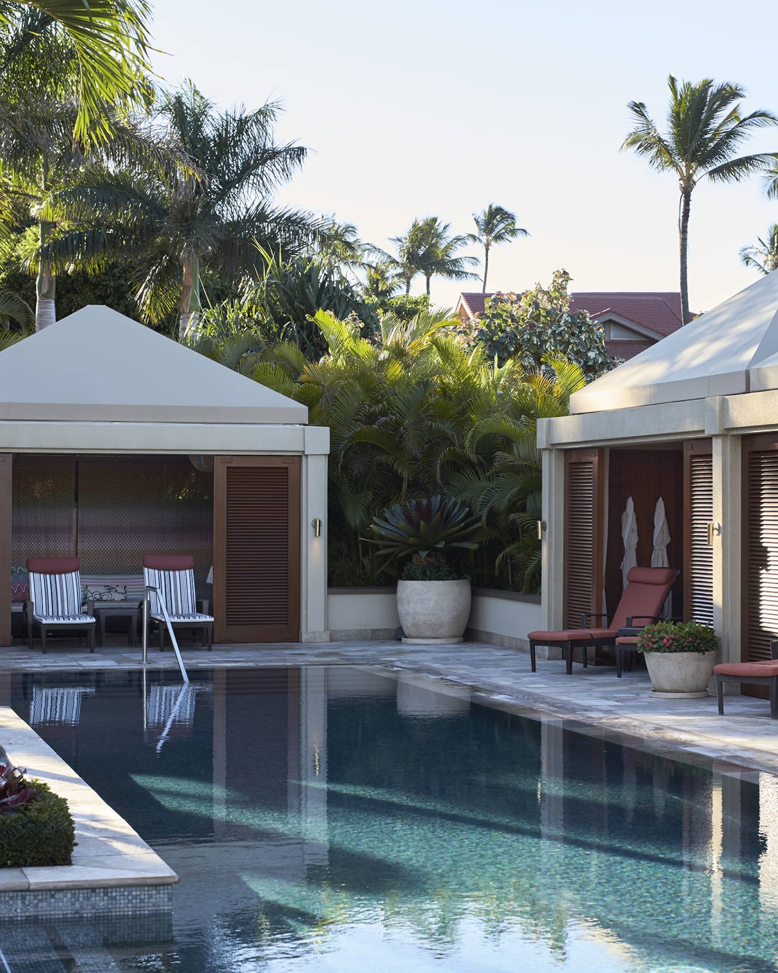 Cabanas line outdoor swimming pool deck under palm trees and sunny sky