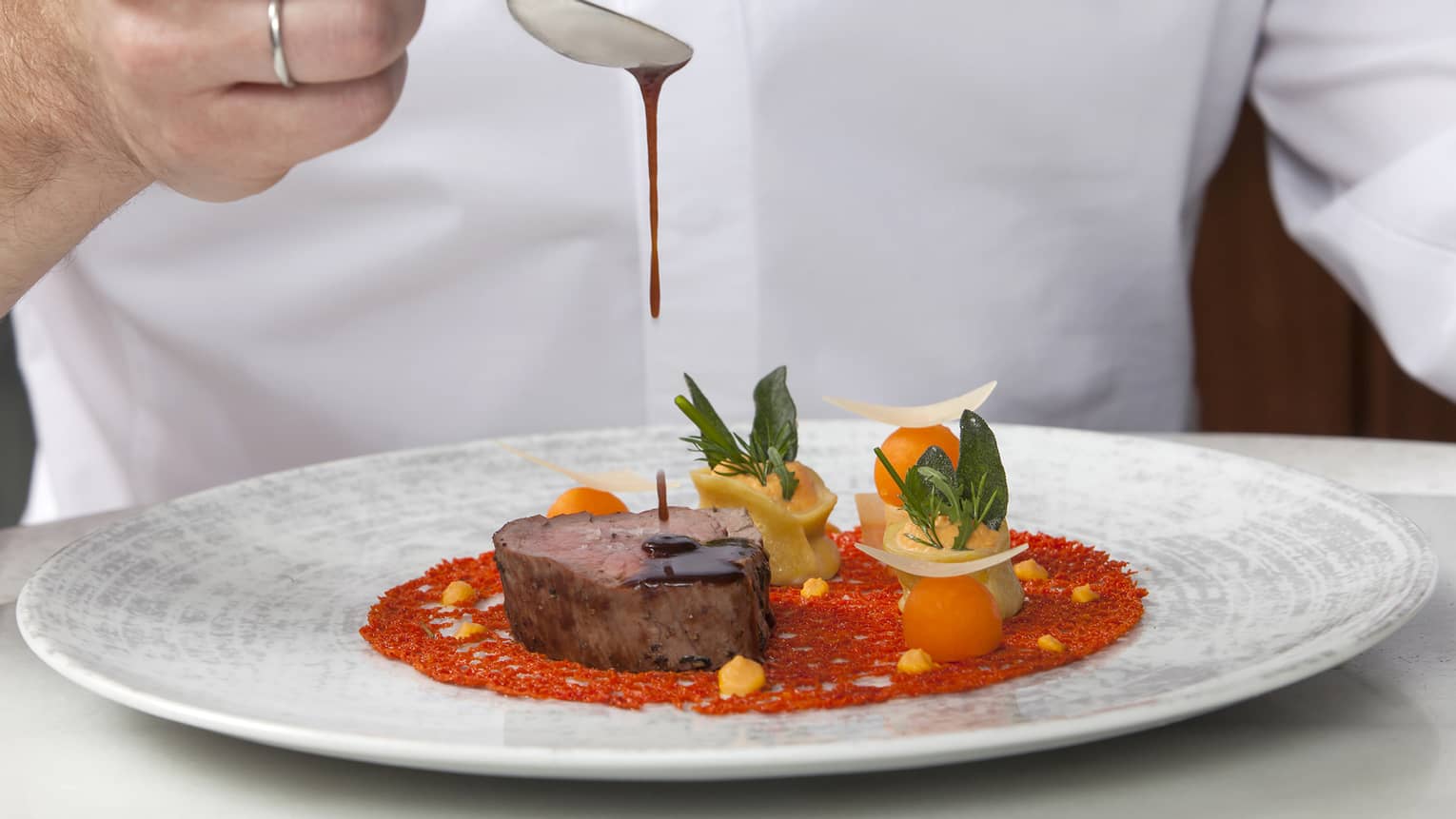 Chef holds spoon over plate, drizzles sauce onto steak entree with artistic plating
