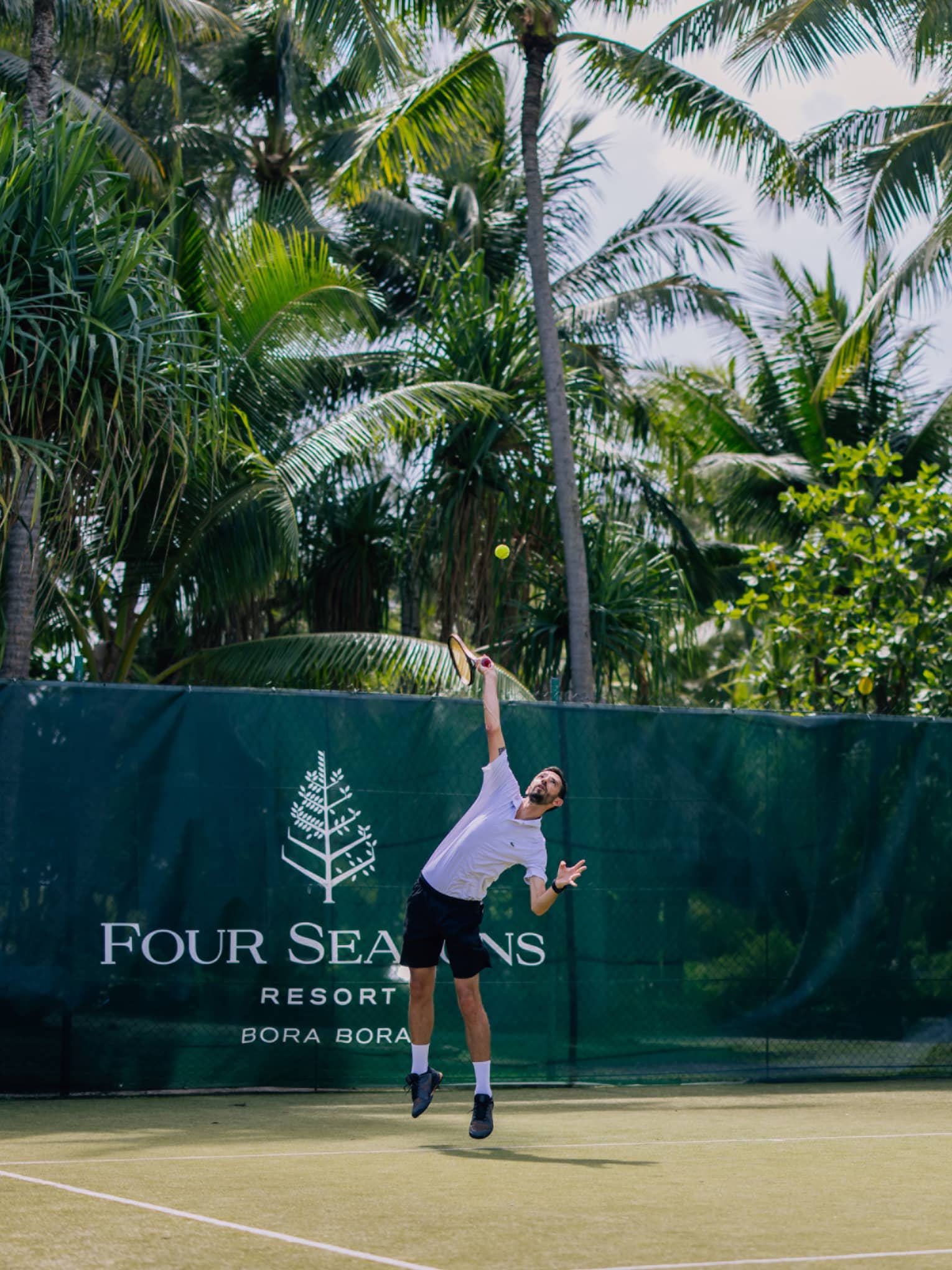 Person in athletic wear, serving on tennis court surrounded by palm trees