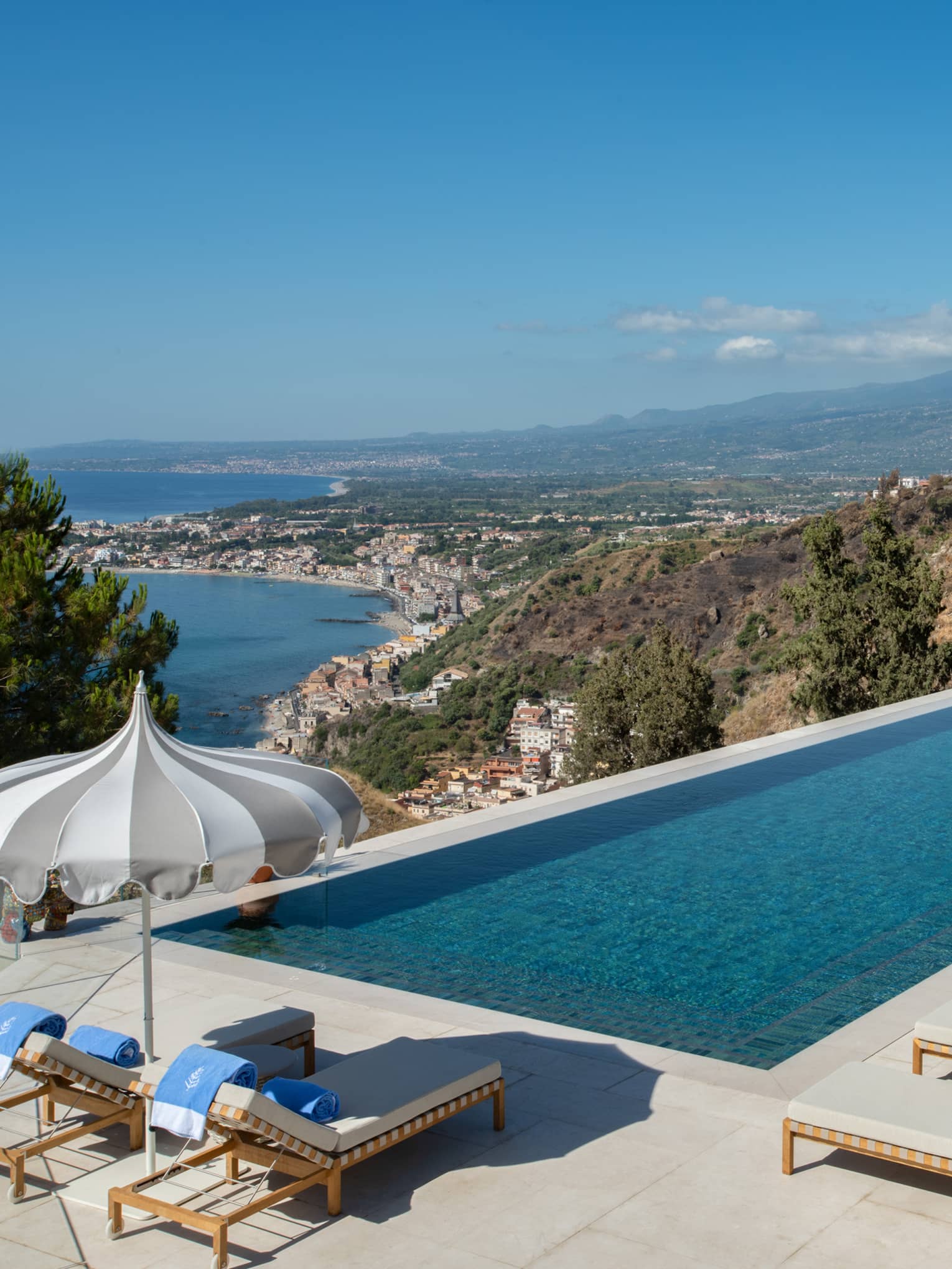 Rectangular pool, lounge chairs and umbrellas overlooking sea and Mount Etna