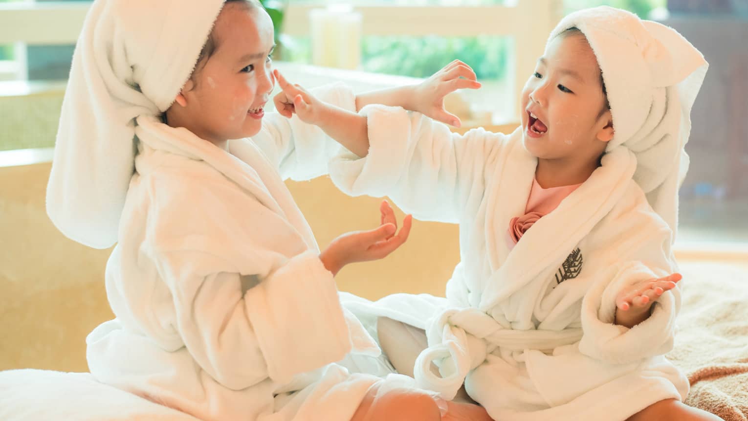 Two laughing young children in white bathrobes with towels around hair put lotion on each other's faces 