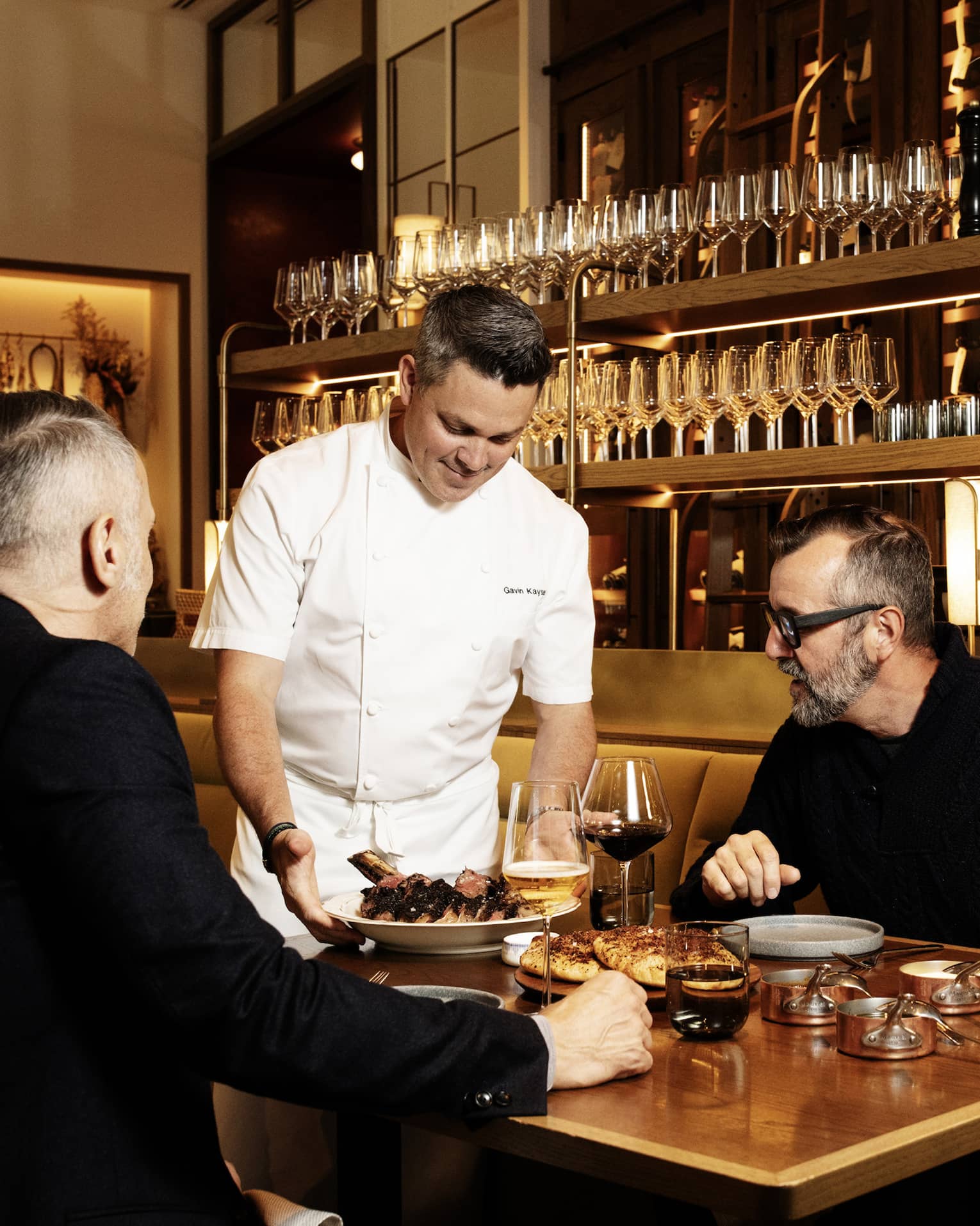 Chef brings a plate of food to two people seated at a table in front of a wood-and-brass shelf of wine glasses