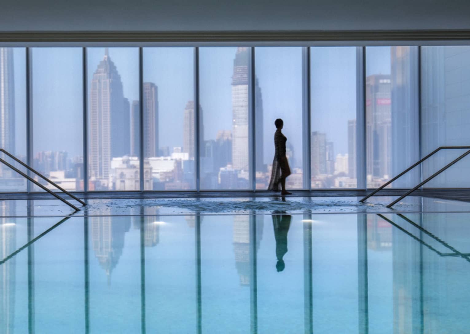 Silhouette of woman walking by blue indoor swimming pool, floor-to-ceiling window