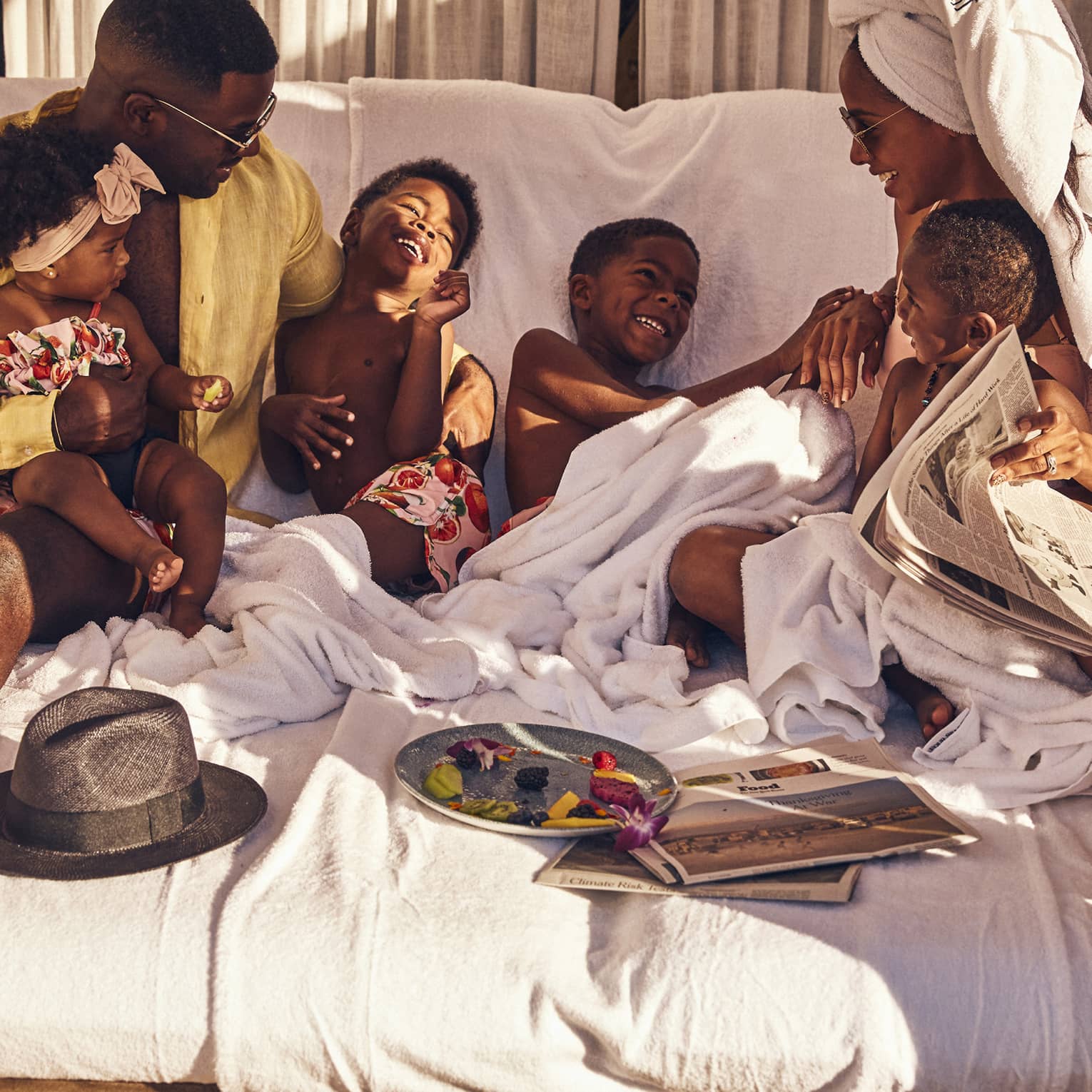 A family of six in bathing suits and towels smile as they play on a cabana bed. A leftover fruit plate sit in front of them.