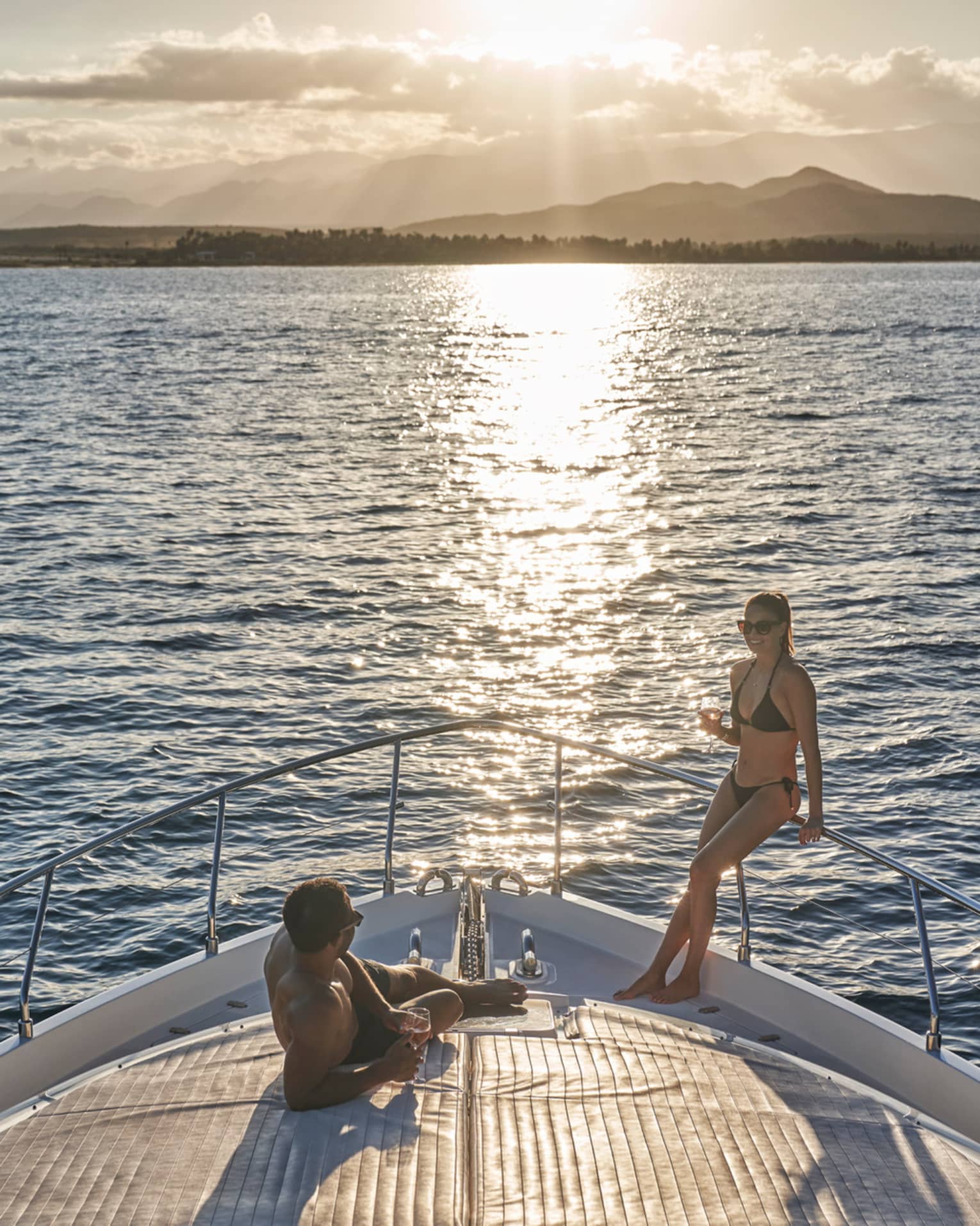 A couple in swimwear relaxing on the bow of a boat, wine glasses in hand and the setting sun glinting off the endless ocean.