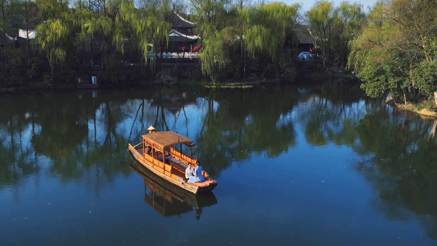 Facing each other, two guests sit in the front of a small wooden boat on a peaceful lake as a third person steers the craft.