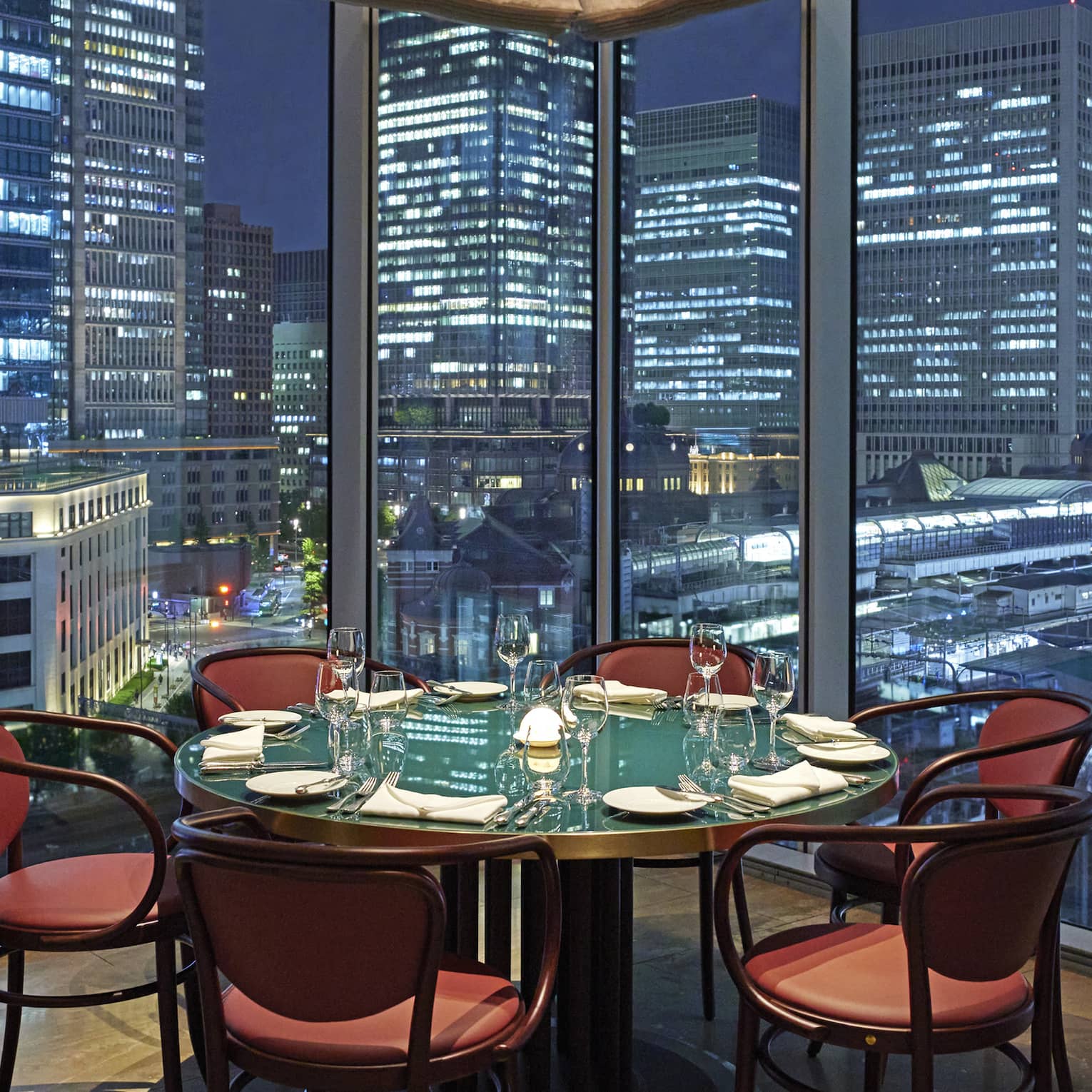 Luxury boutique hotel dining setup with a round table elegantly set for guests, offering a panoramic city view at night through tall glass windows