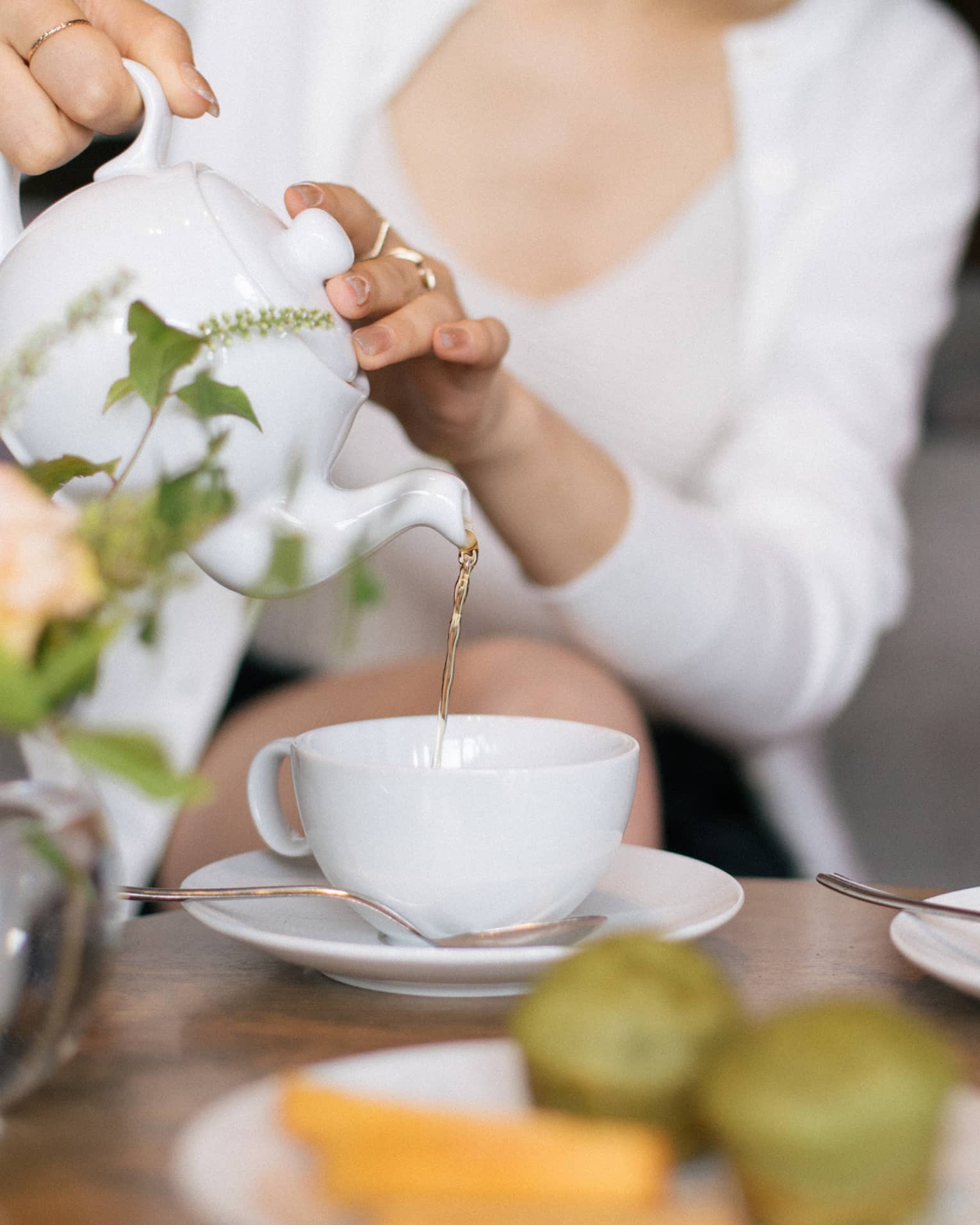 Two people sit at a table set for tea, with cups, saucers, a plate of small food items and a floral arrangement. One person is pouring tea from a teapot into one of the teacups.