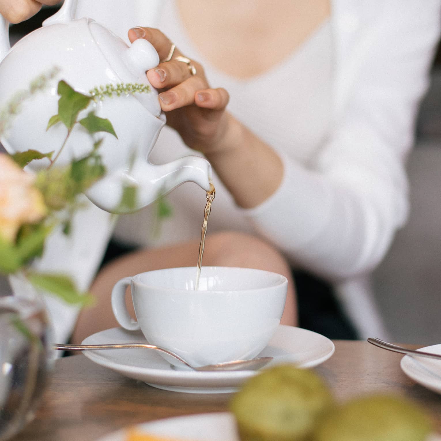 Two people sit at a table set for tea, with cups, saucers, a plate of small food items and a floral arrangement. One person is pouring tea from a teapot into one of the teacups.
