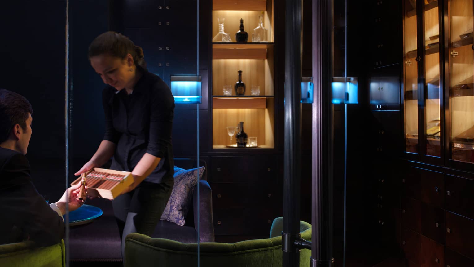 Woman holds open box of cigars, displays them to man sitting in dark lounge