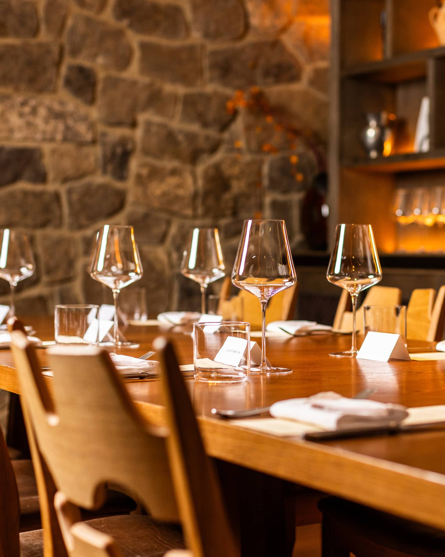 Table set for six in a dining room with a stone walls and a shelf filled with wine glasses