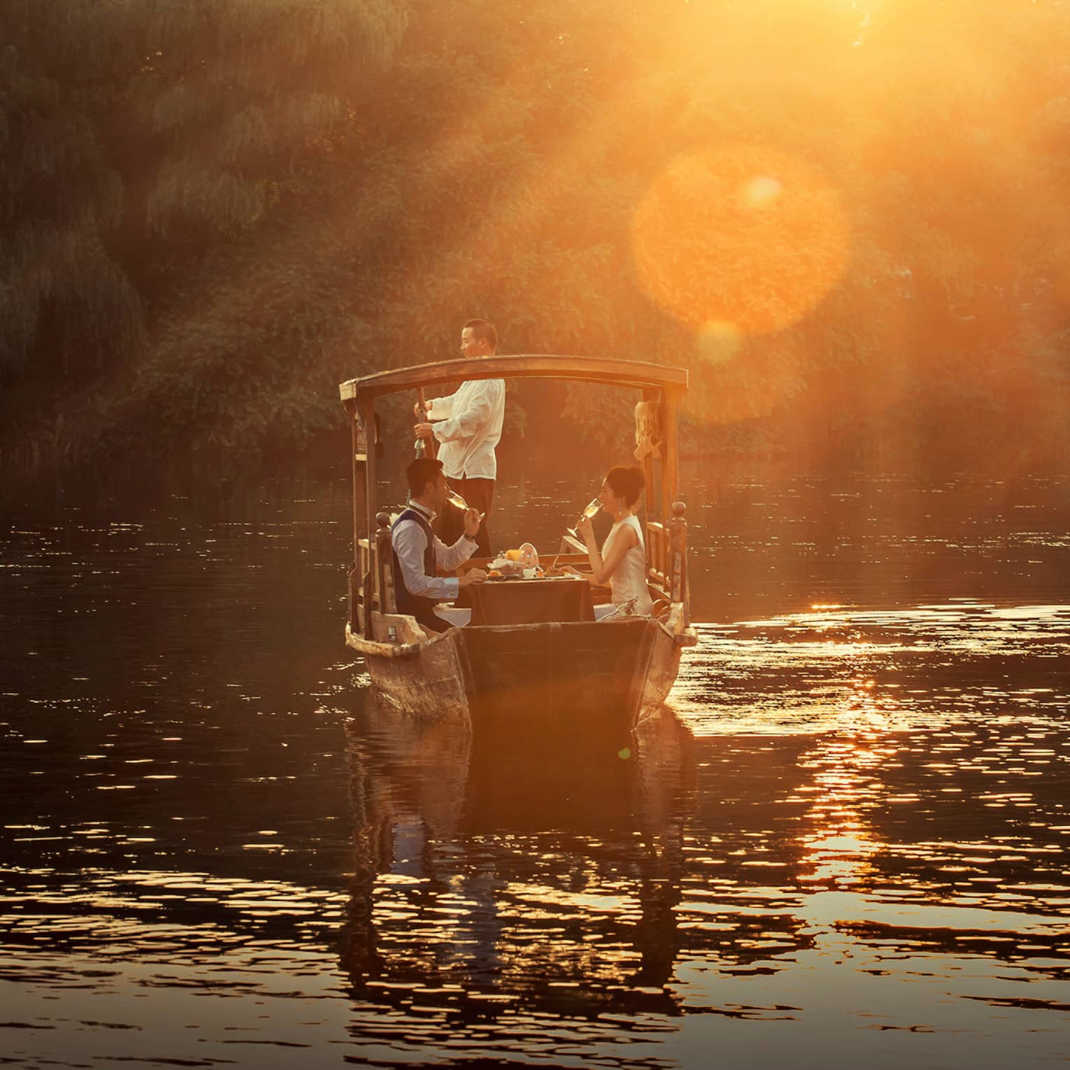 View from behind of a rustic wooden boat on a calm pond, under a golden glow; a couple enjoys champagne at a table inside.
