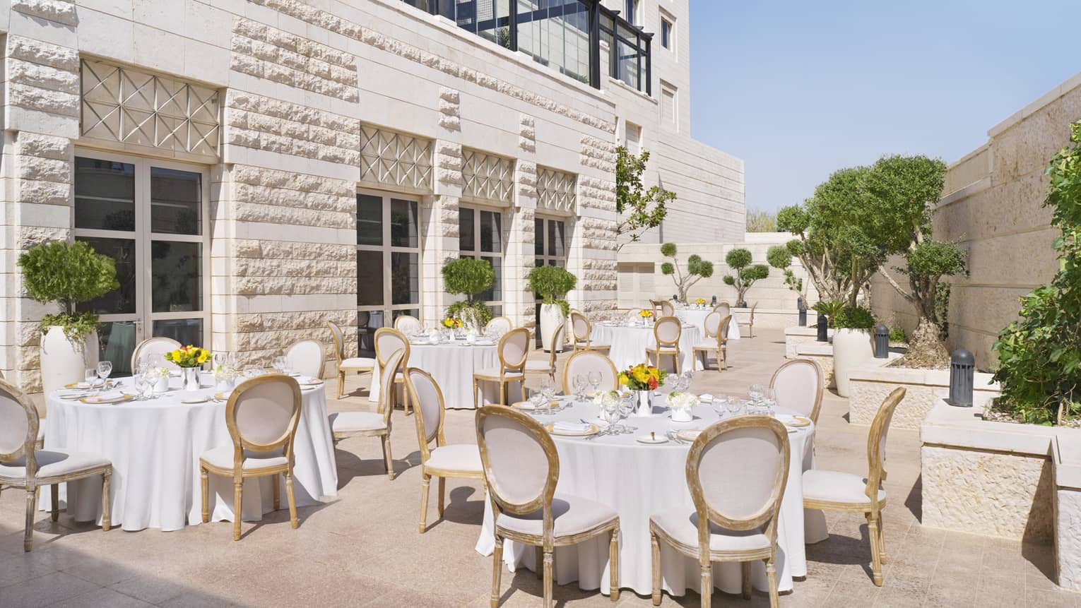 A series of circular tables covered in white linen, and surrounded by gold chairs on a patio.