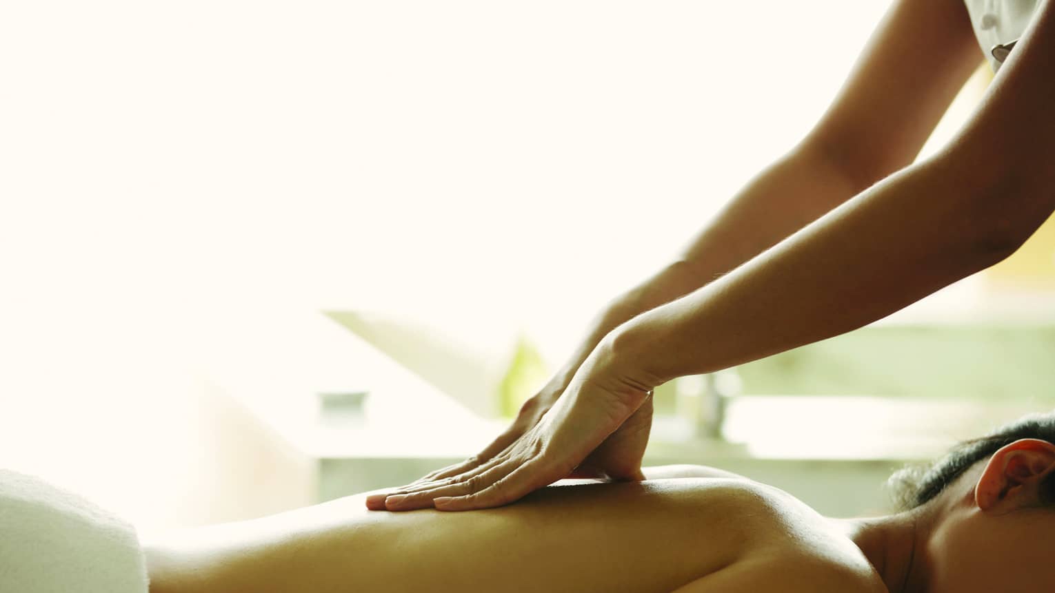 Masseuse reaches over, massages woman's bare back as she lies on spa table