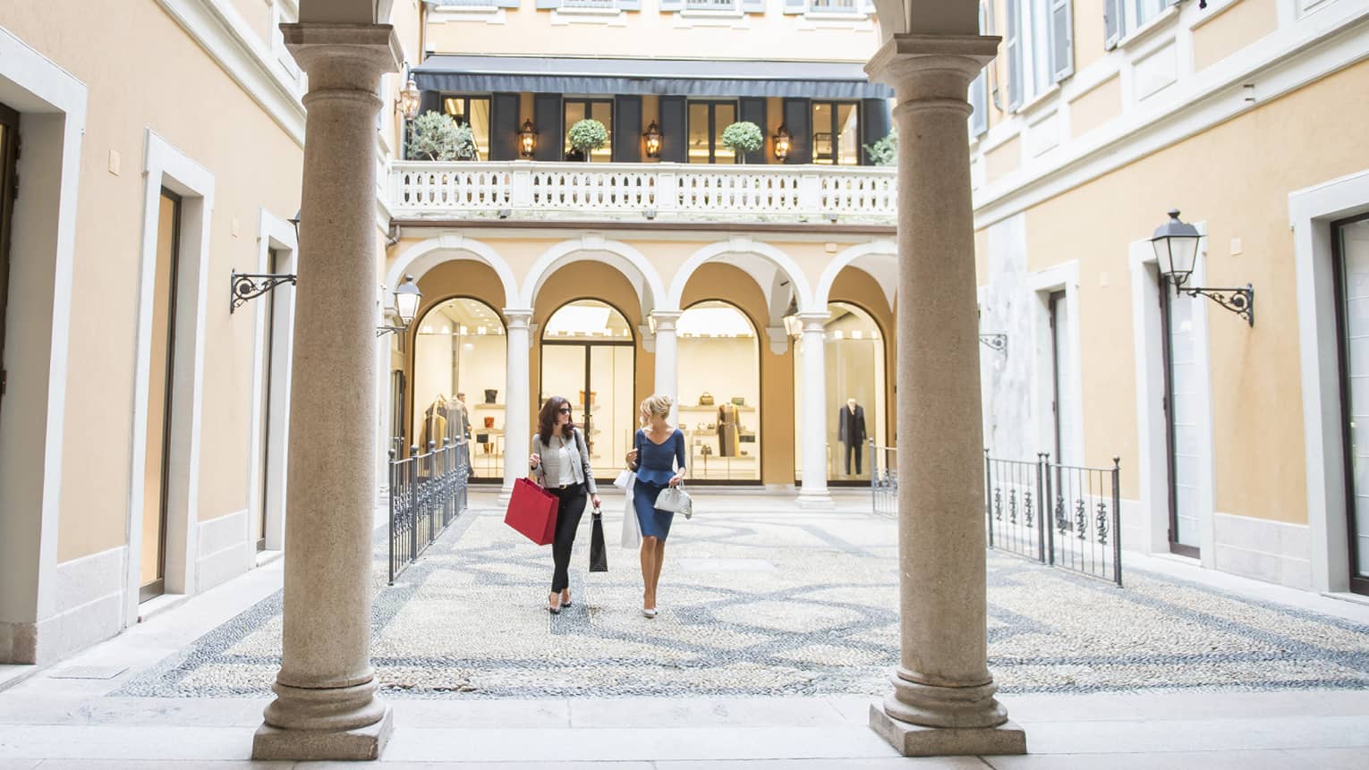 Two women with shopping bags walking through a sunlit courtyard