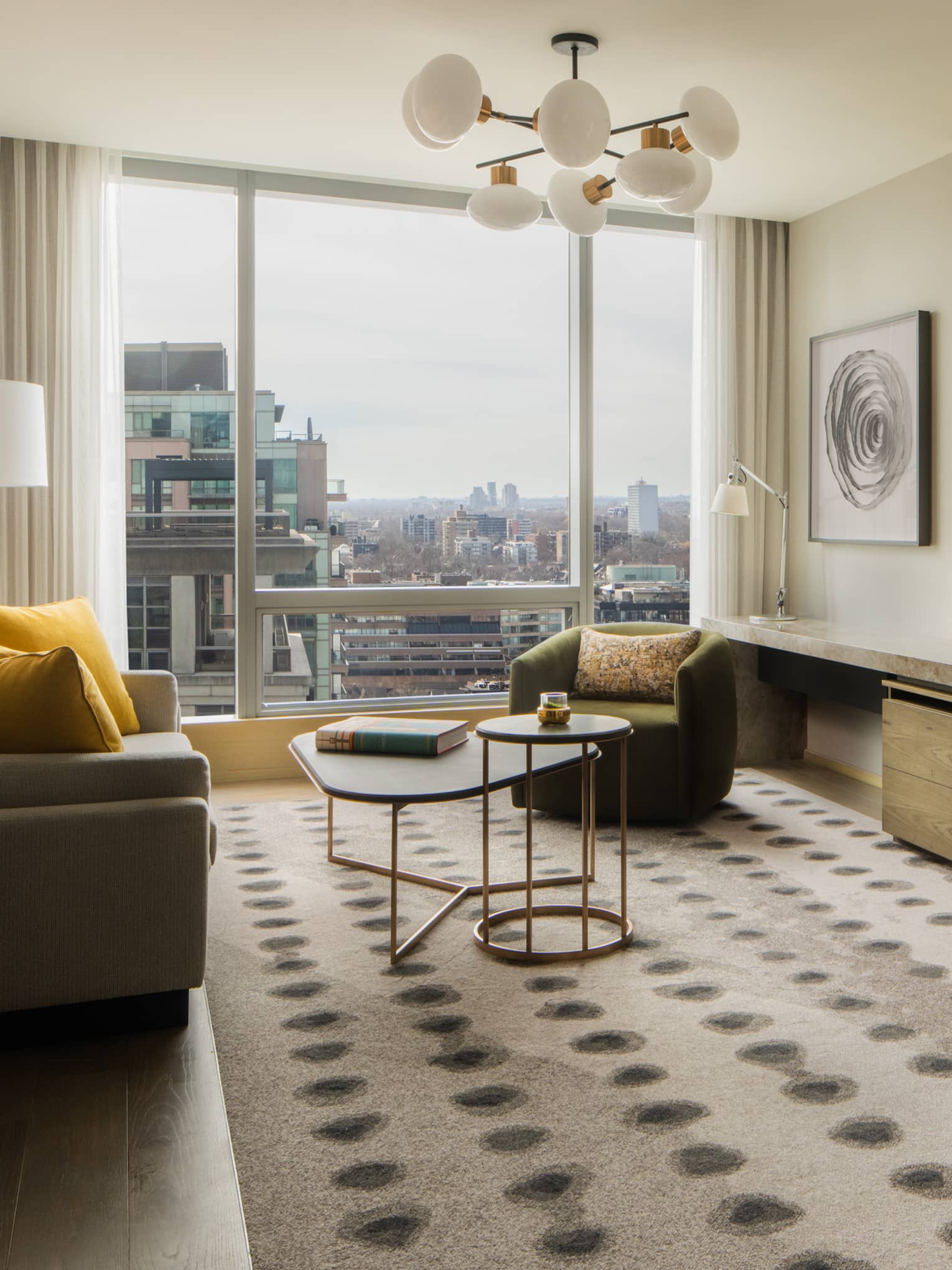 Hotel suite living room with a beige sofa, yellow pillows, armchairs, coffee tables, wall-mounted TV, console table, abstract artwork and large windows