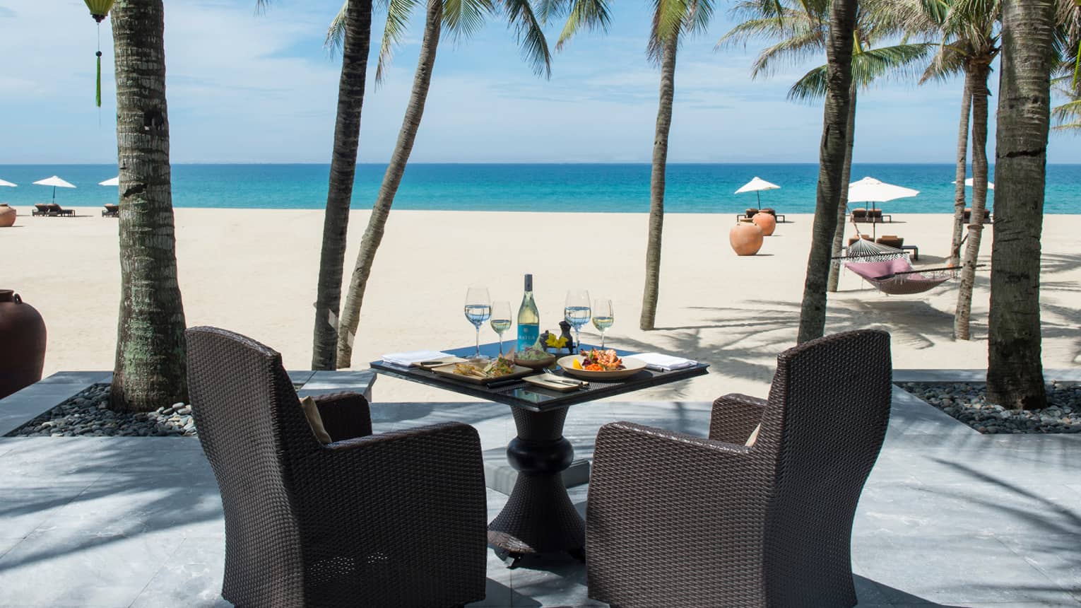 Dining table with meals, white wine, large black wicker chairs facing beach, ocean