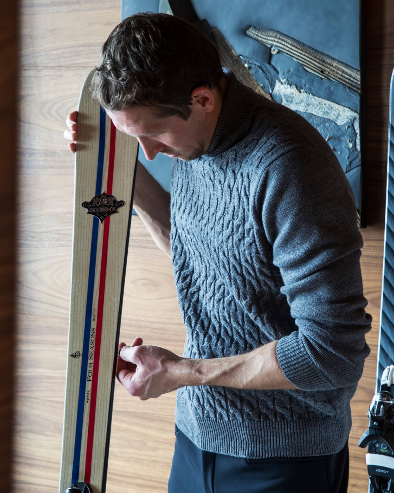 Man wearing knit sweater leans over ski against wall, more skis on wall